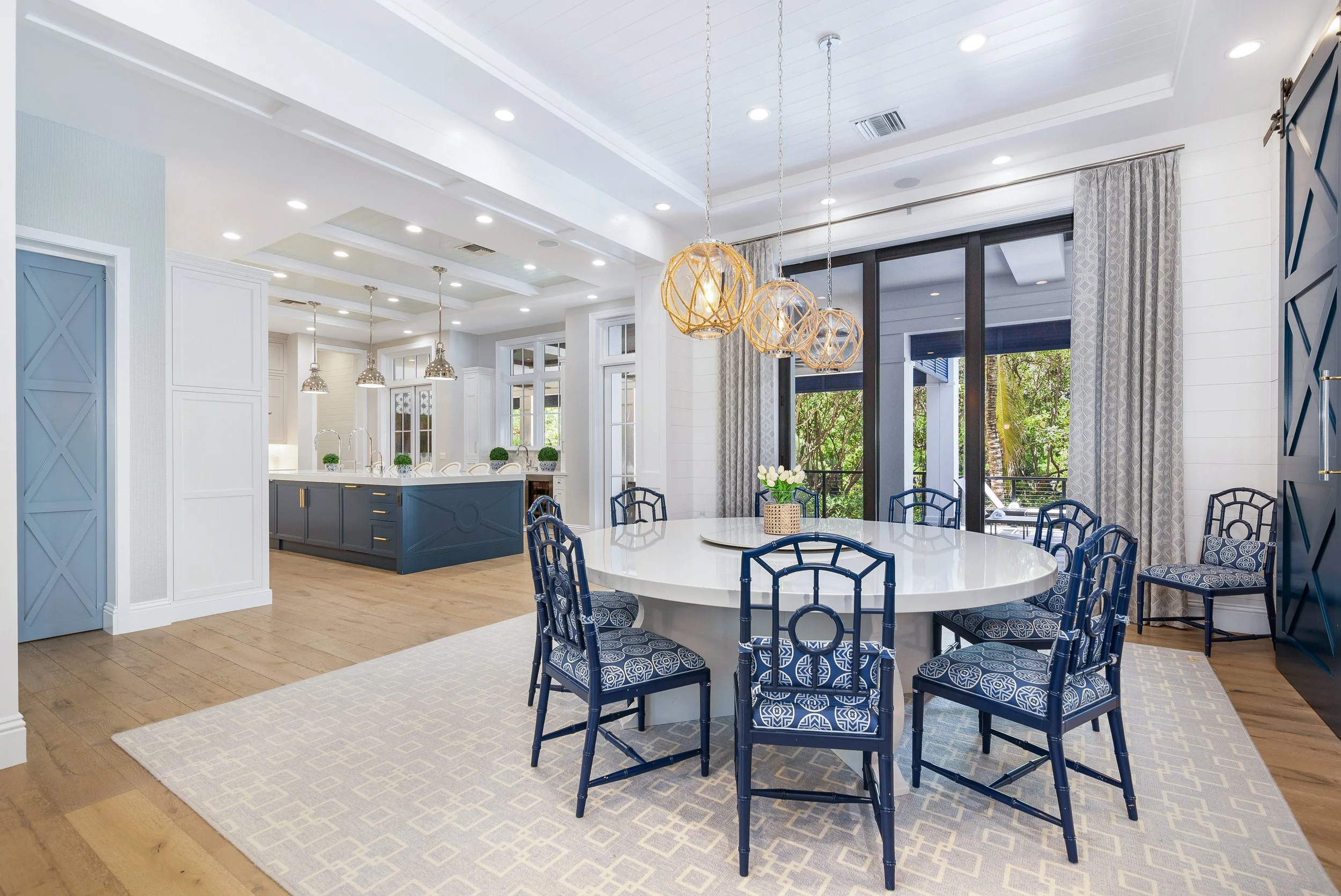A bright, modern open-concept dining and kitchen area with large windows, white walls, and a high ceiling. The dining table is white with blue chairs, and a flower centerpiece. The kitchen island is blue with a white countertop. Hanging pendant light