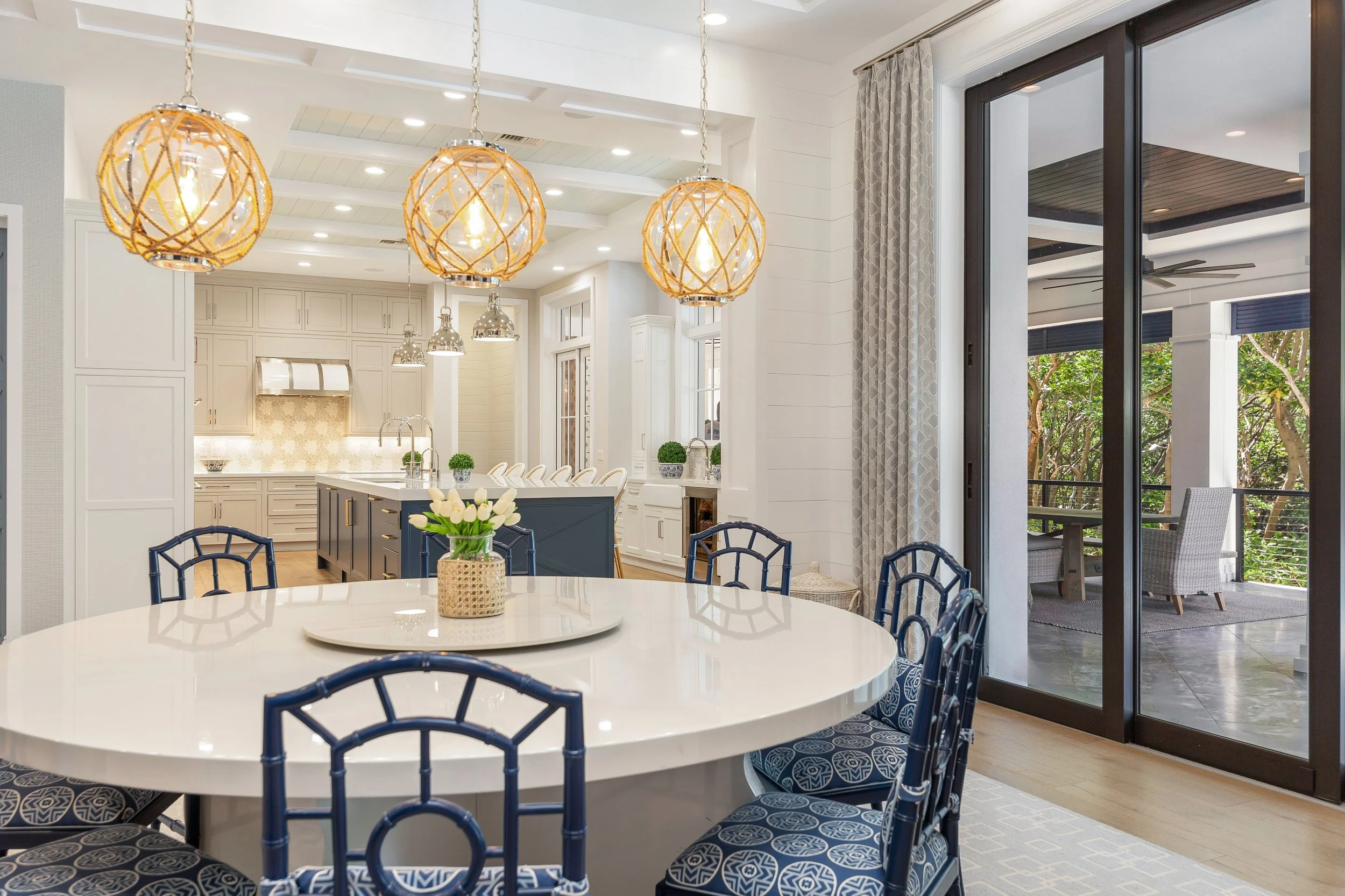 A bright, modern kitchen and dining area with pendant lights over a large white round table. The table has a small vase with white flowers and surrounded by navy blue chairs with patterned cushions. There is a sliding glass door leading to an outdoor