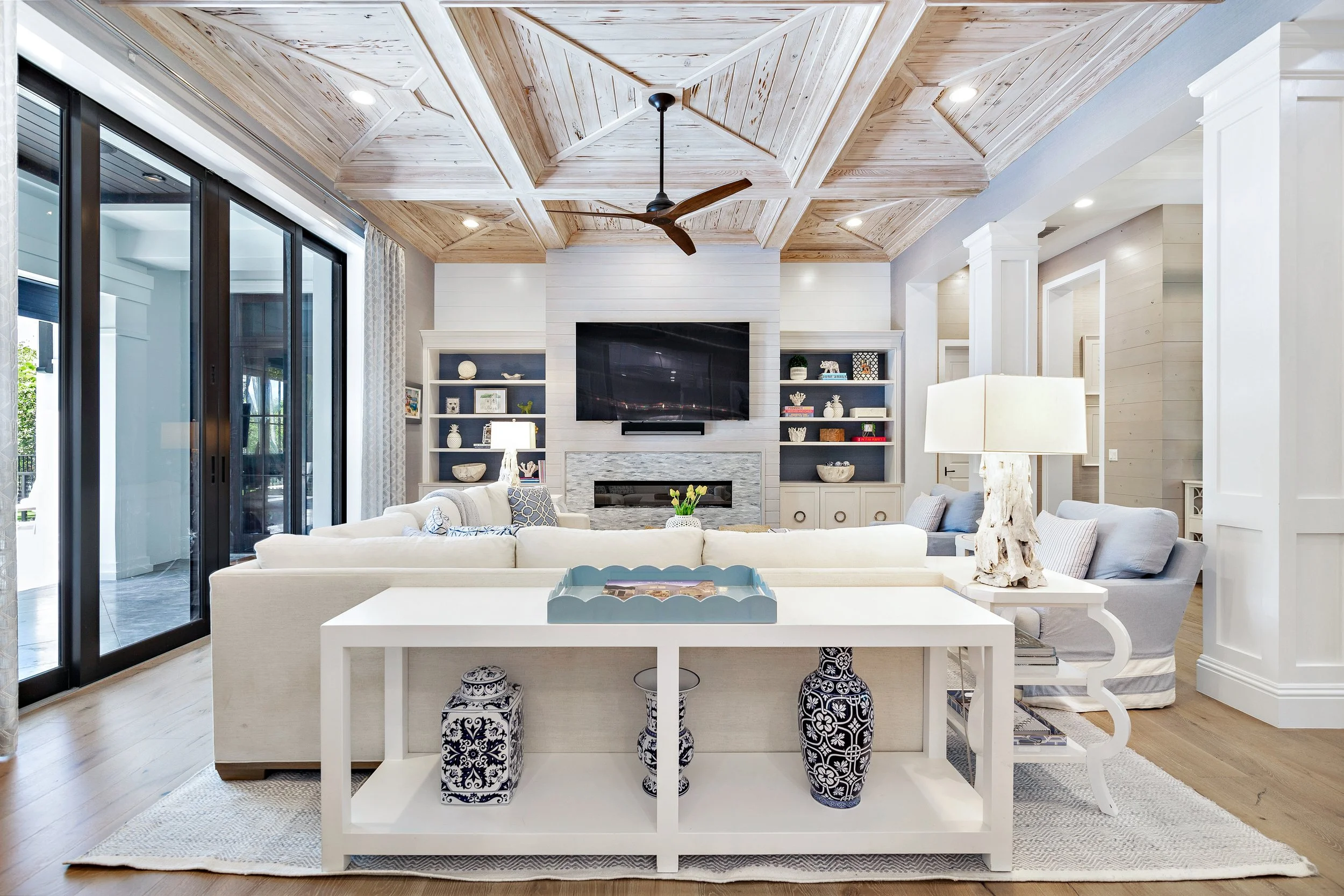 Bright living room with a wooden coffered ceiling, a large black flat-screen TV, white built-in shelving units with decorative items, a light gray sofa, a white coffee table, and tall sliding glass doors with curtains leading outside.