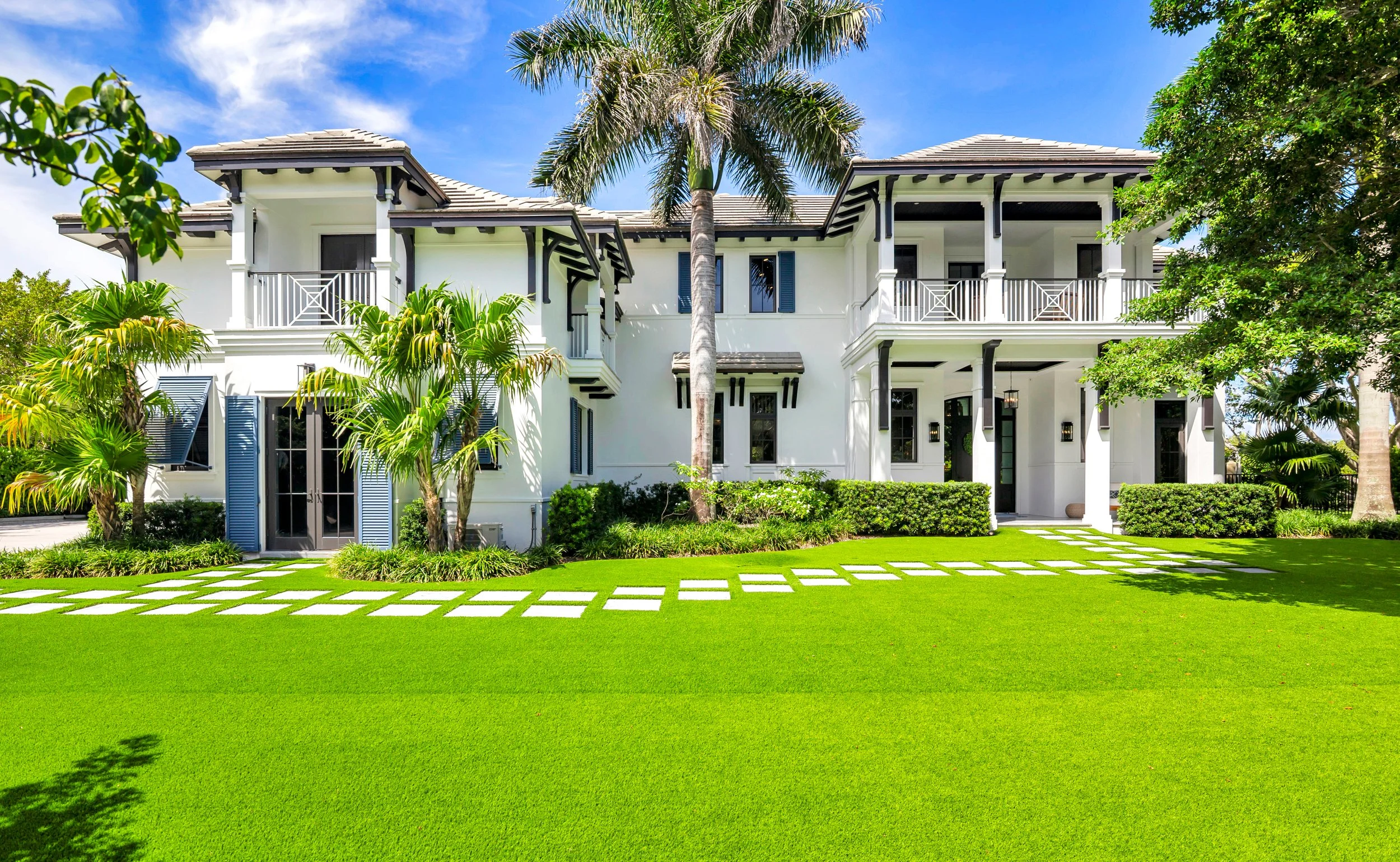 A large white house with black accents, multiple balconies, and a variety of windows, surrounded by lush green trees and a well-maintained lawn with a stone pathway.