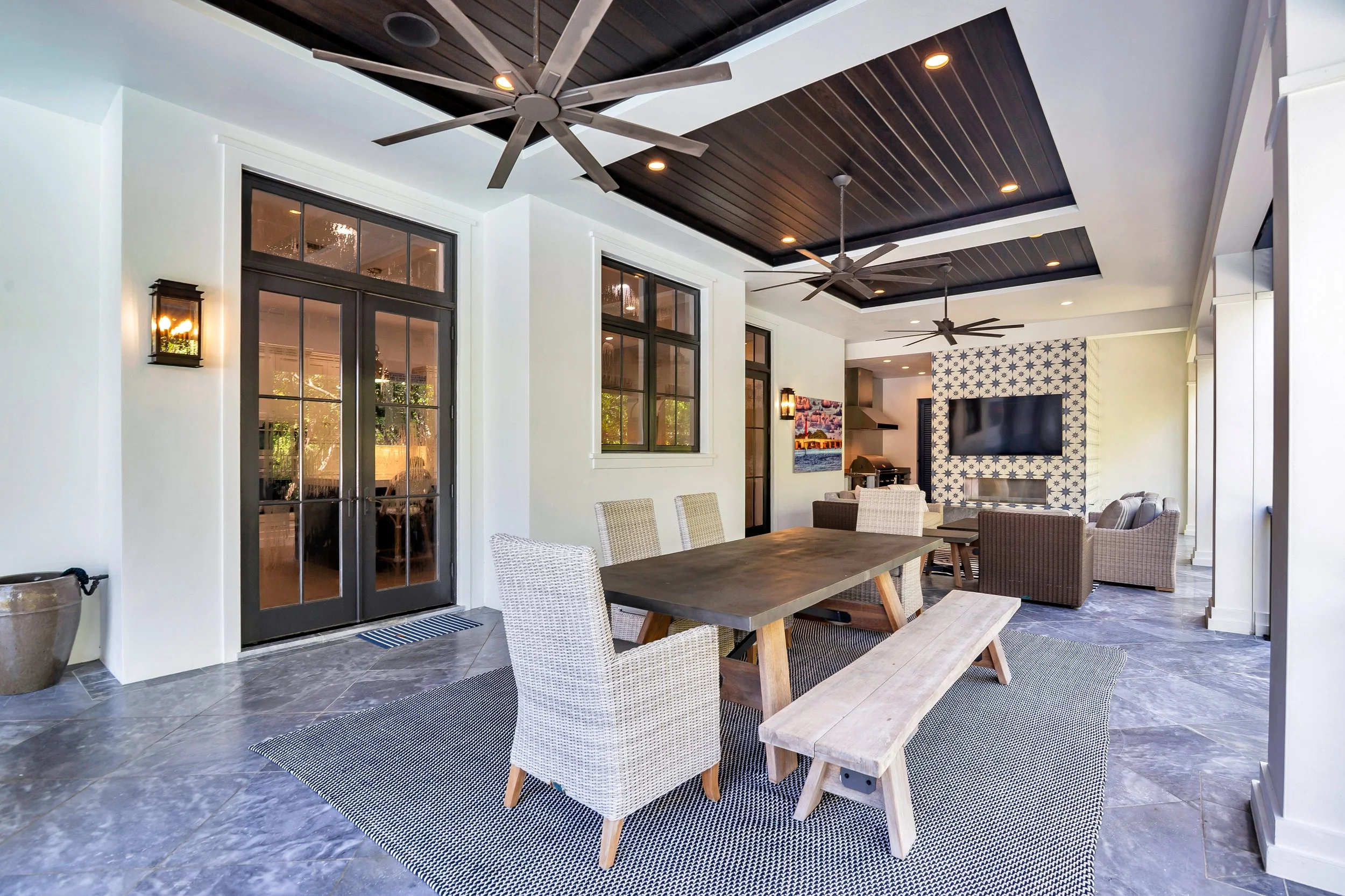 A covered outdoor living space with ceiling fans, recessed lighting, large windows, and a TV mounted on a patterned wall. There is a dining table with chairs and a small bench, and the floor is tiled.