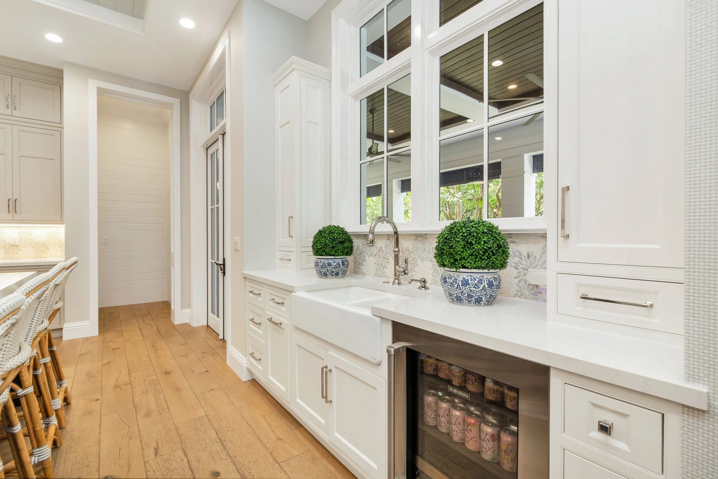 Kitchen with white cabinets, a granite countertop with two decorative potted plants, a wine fridge stocked with cans, large windows with a view of greenery outside, hardwood flooring, and a ceiling with recessed lighting.