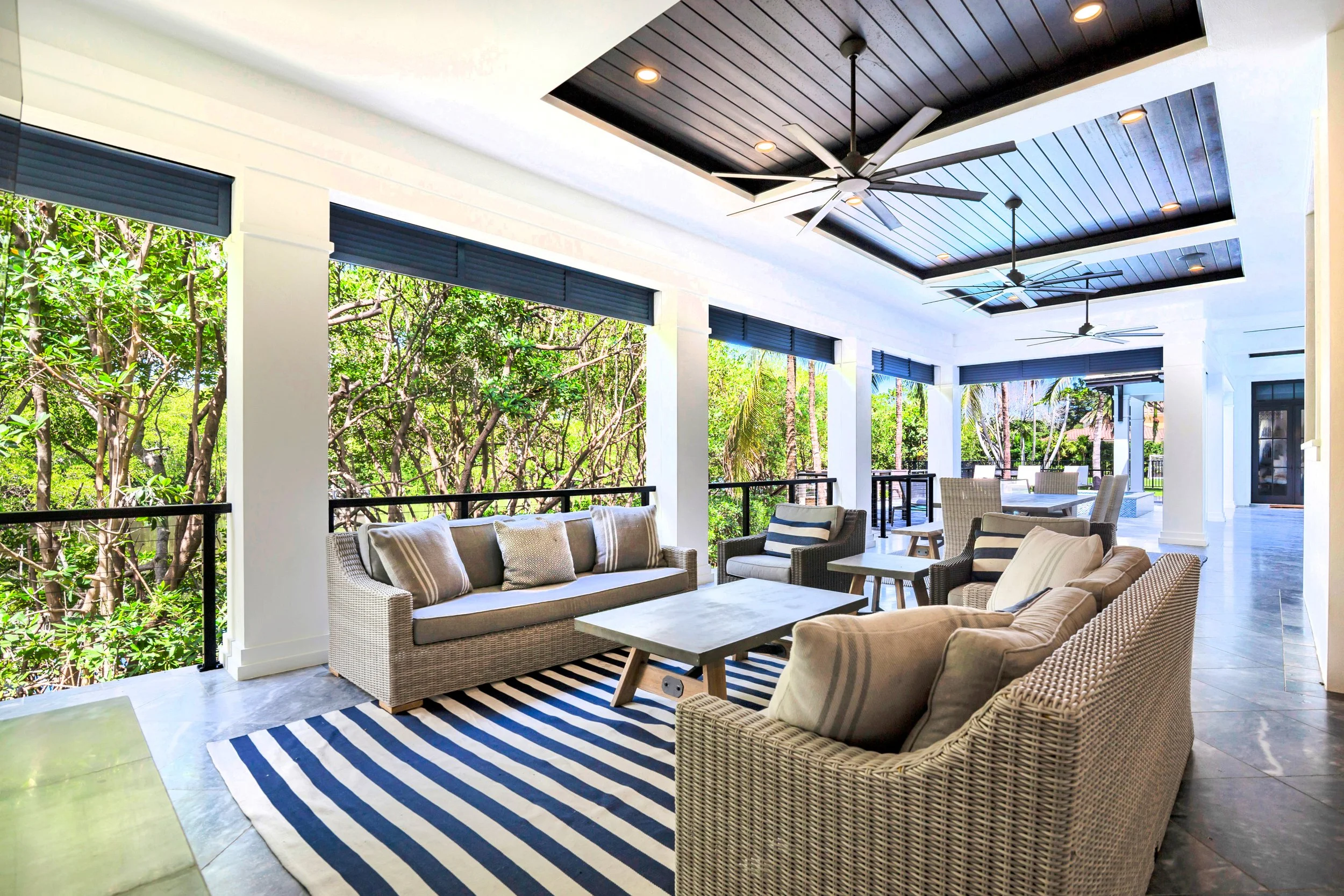 Covered outdoor patio area with wicker furniture, cushions, and a striped rug, overlooking lush greenery and trees, with ceiling fans and recessed lighting in a modern home.