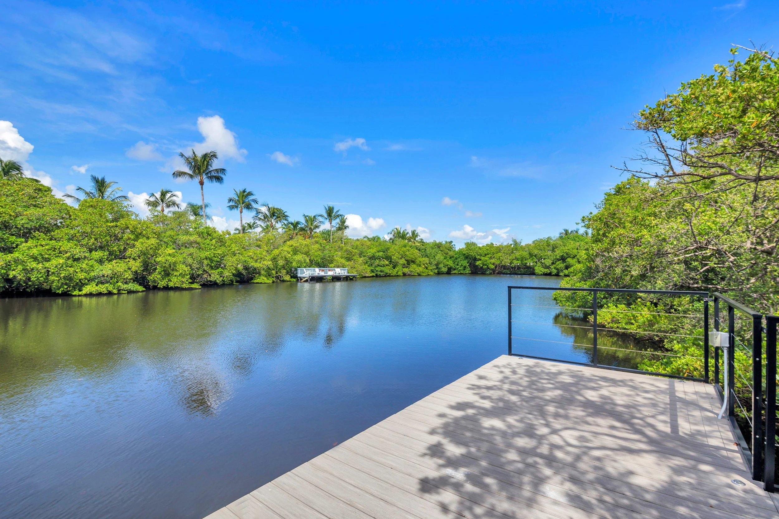 A scenic view of a river with lush green trees on both sides under a bright blue sky with a few clouds. A wooden deck with a metal railing extends into the water in the foreground.