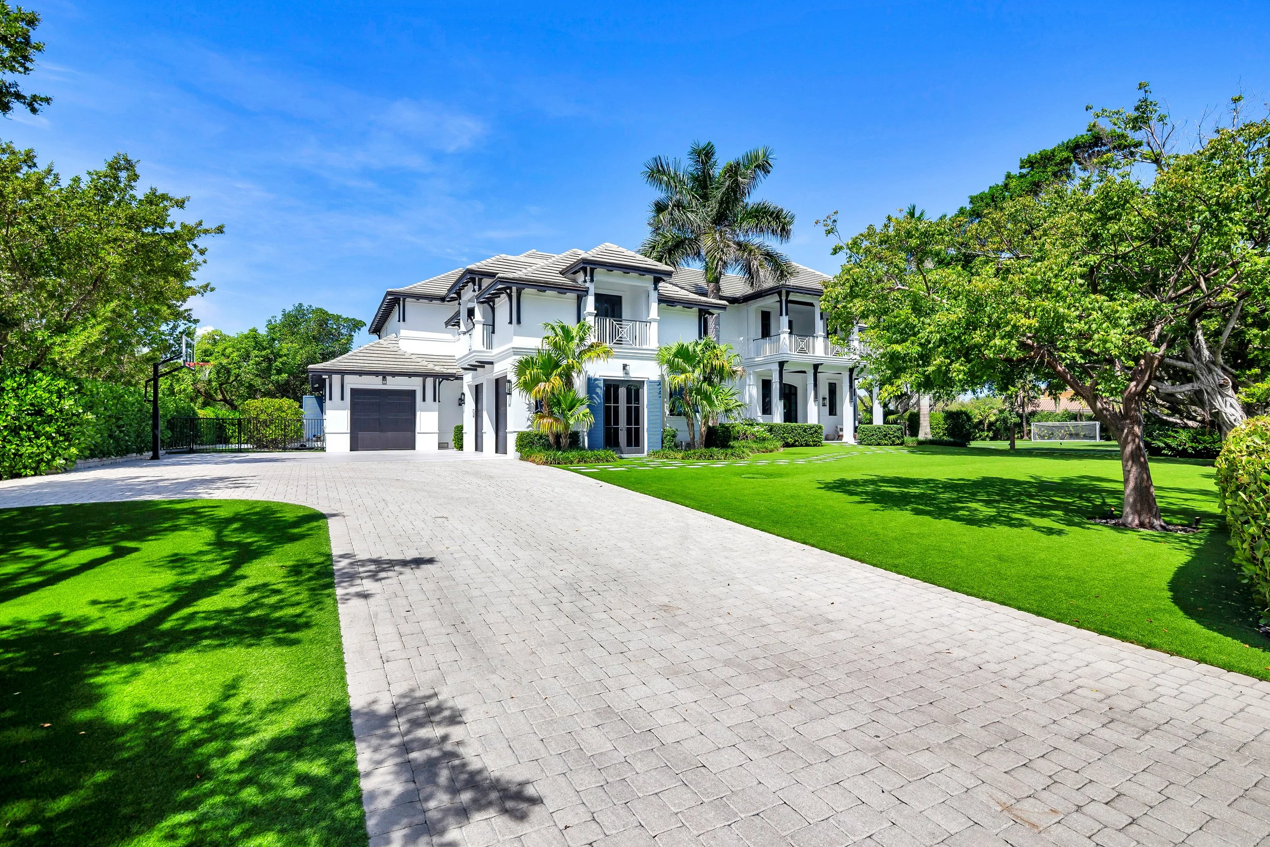 A white two-story house with black accents, surrounded by lush green lawns and trees, with a paved driveway and clear blue sky.
