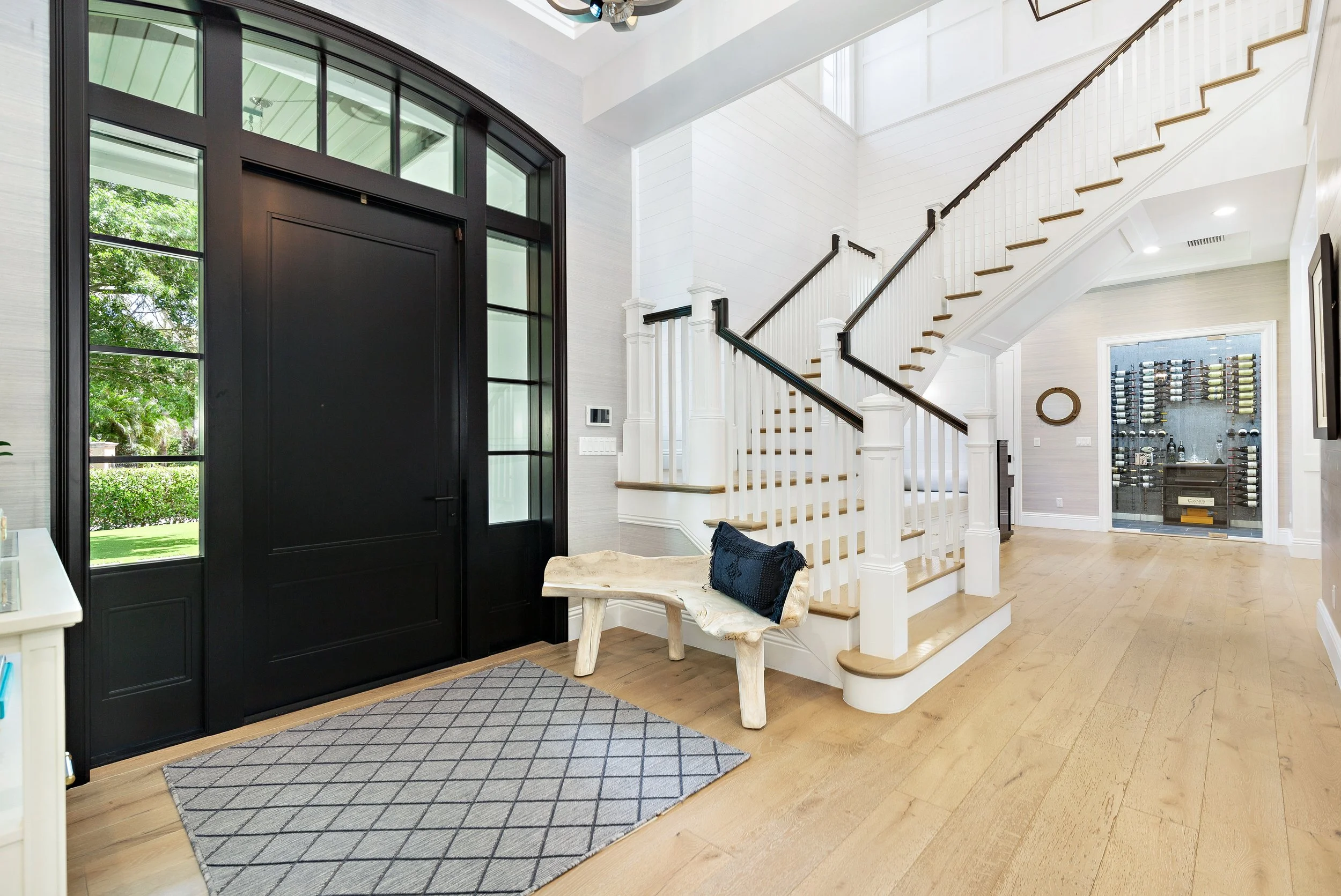 Interior of house entryway with black front door, staircase with white balusters and dark handrail, wine cellar in the background, and seating area with a bench and decorative cushion.