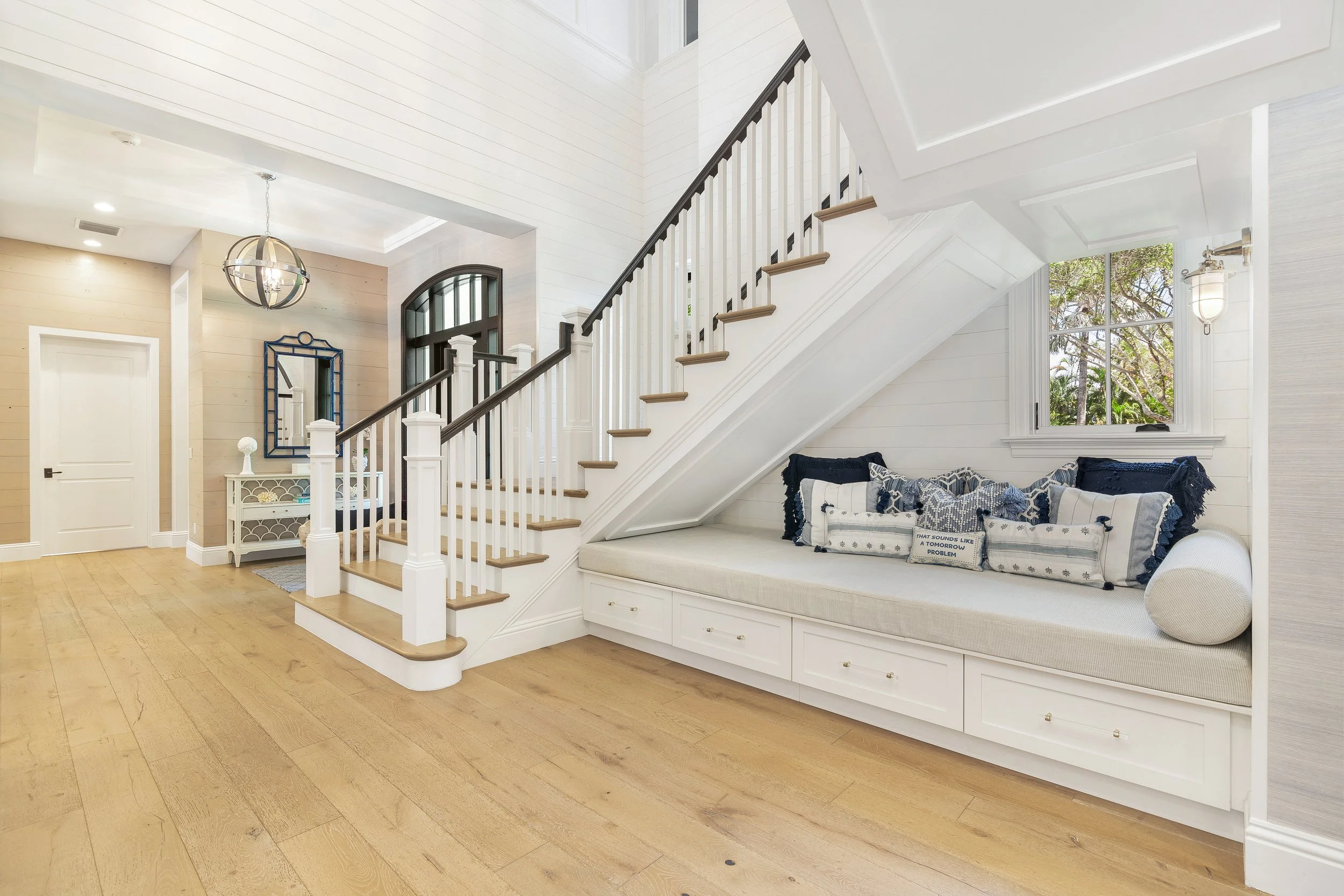 Interior view of a bright, modern living space with a staircase, beige walls, a built-in window seat with blue pillows, a large window showing trees outside, and a decorative mirror on the wall.