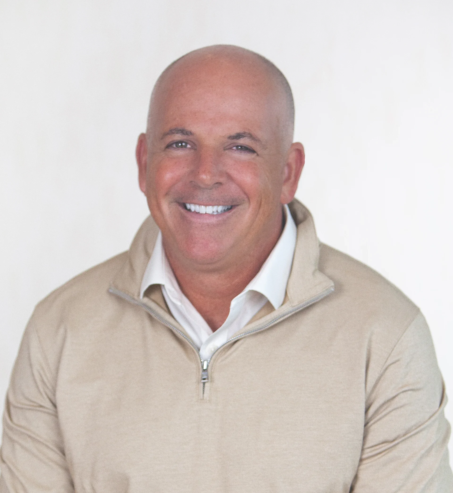 A man with a big smile, bald head, wearing a beige zip-up sweater over a white collared shirt, against a plain white background.