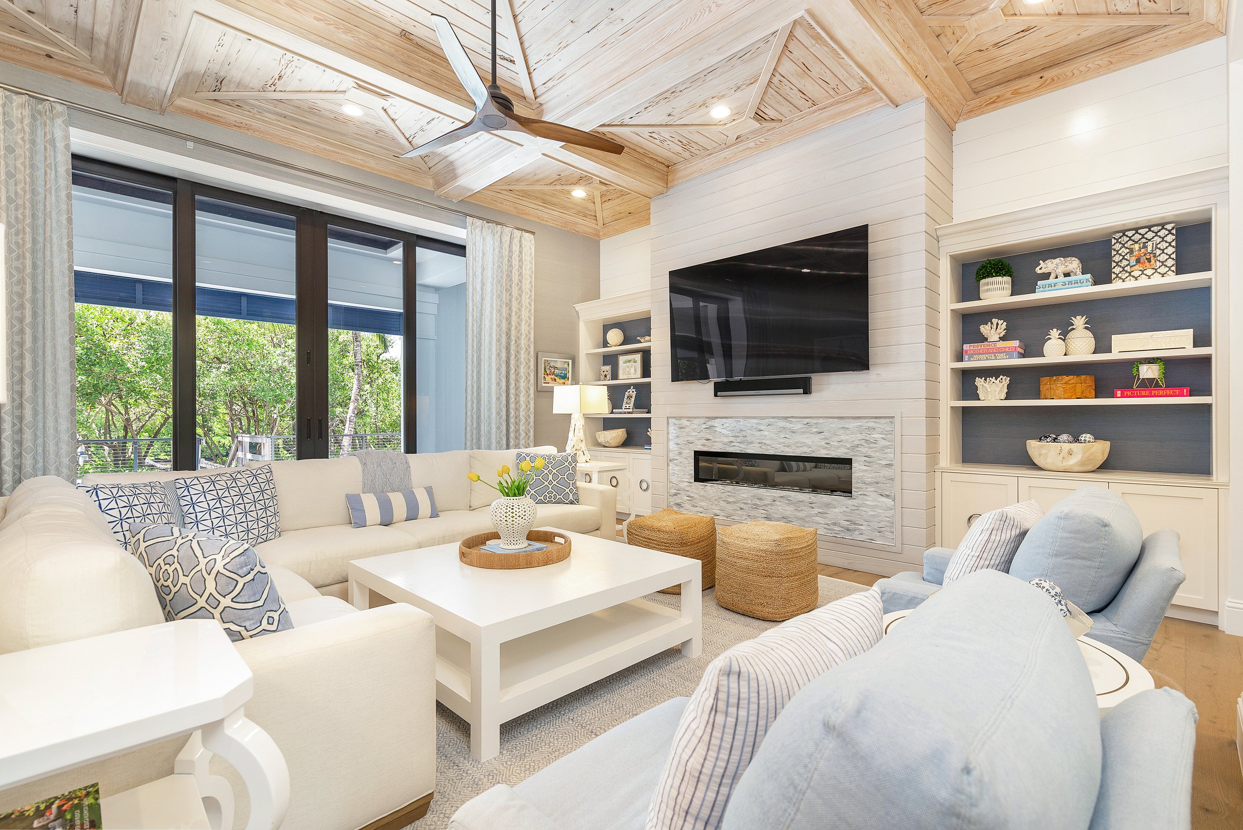 Living room with a vaulted wood ceiling, ceiling fan, large window with trees outside, white sofa with patterned pillows, coffee table, fireplace, and built-in bookshelf with decorative items.