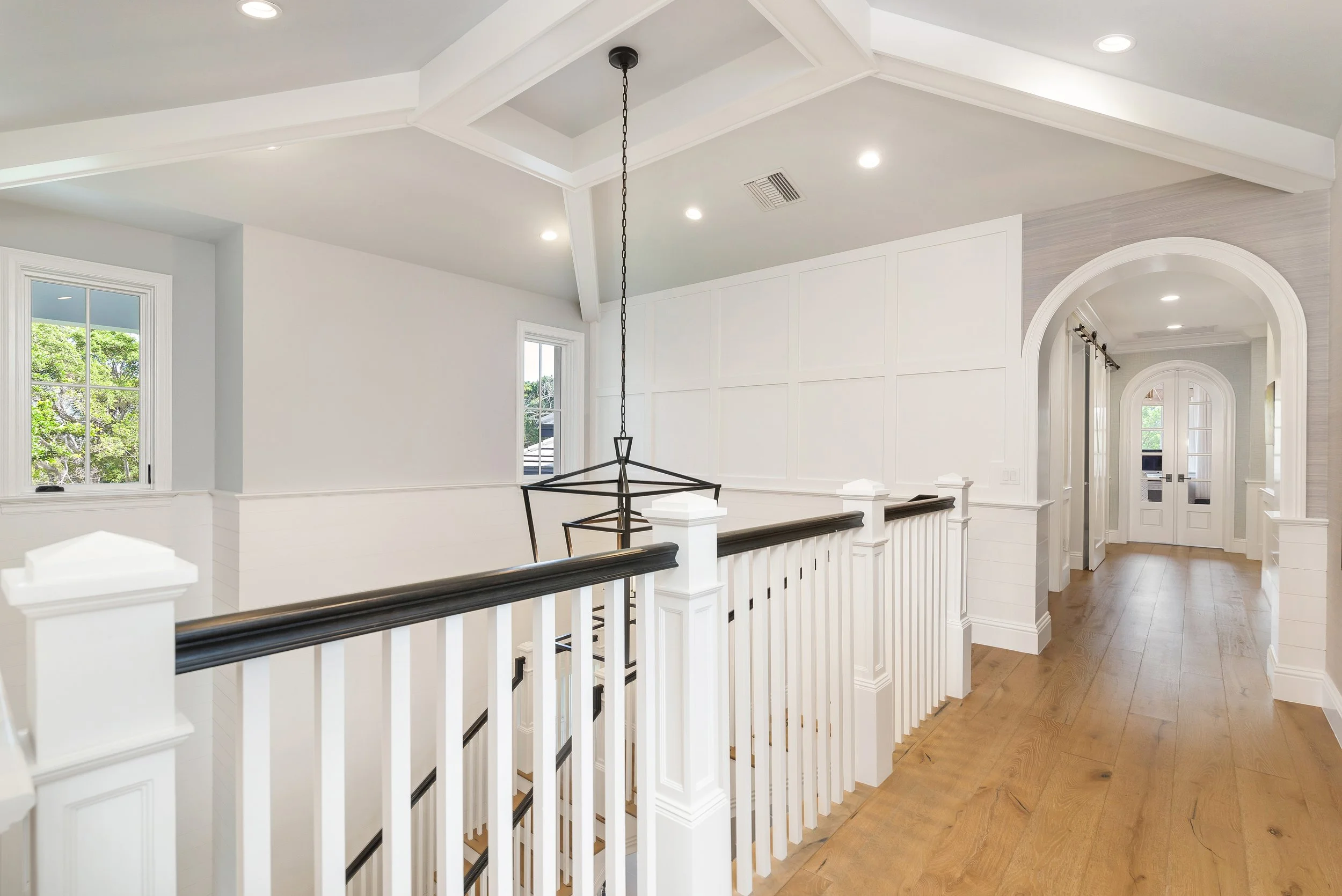 Interior view of a bright, modern hallway with white walls, vaulted ceiling with wood beams, windows letting in natural light, and a staircase with white balusters and dark handrail.