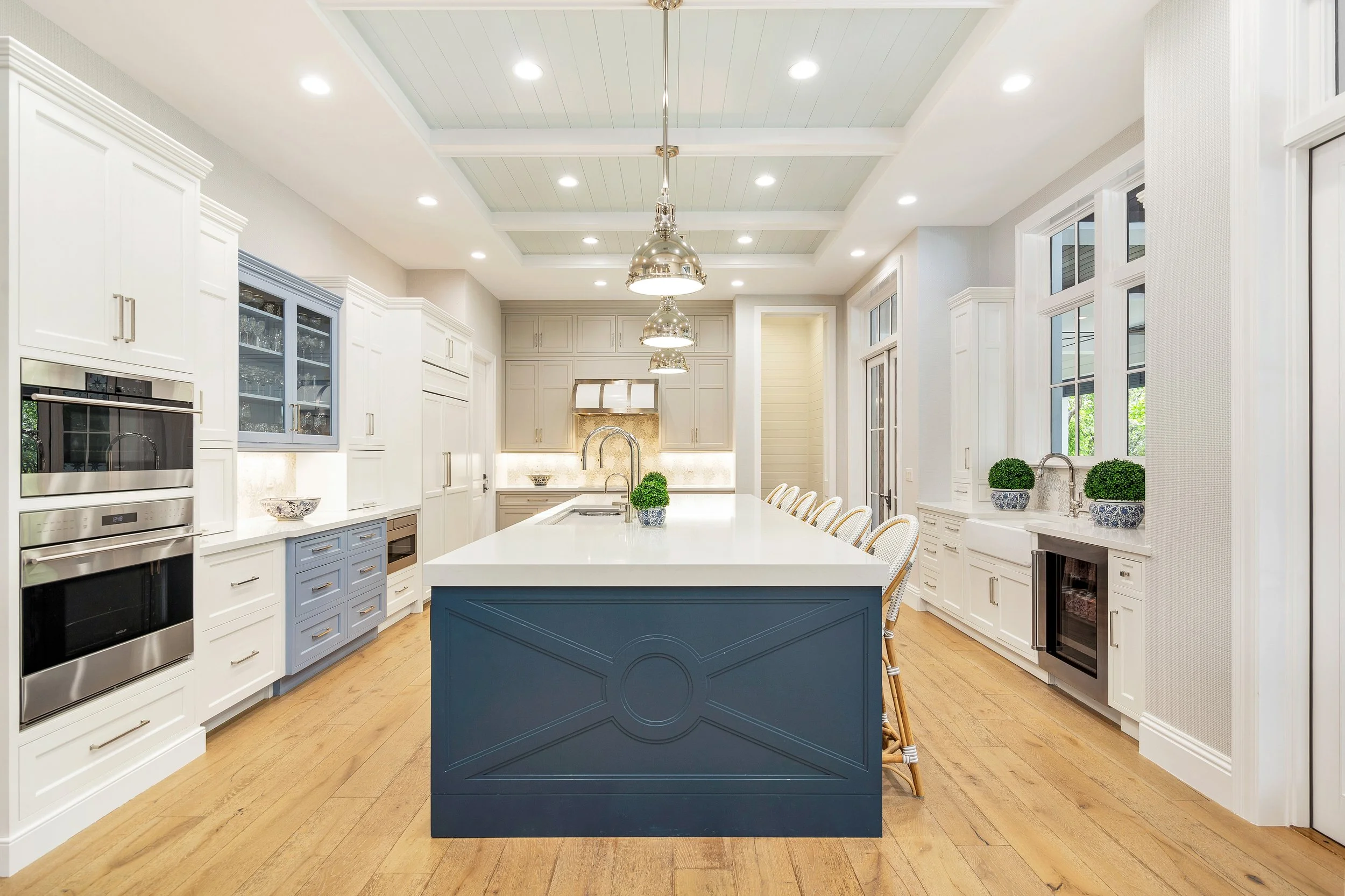 A bright open kitchen with a large white island, blue cabinets, and white cabinets lining the walls. There are three metallic pendant lights hanging over the island, large windows along one wall, and decorative pots with green plants on the counter.