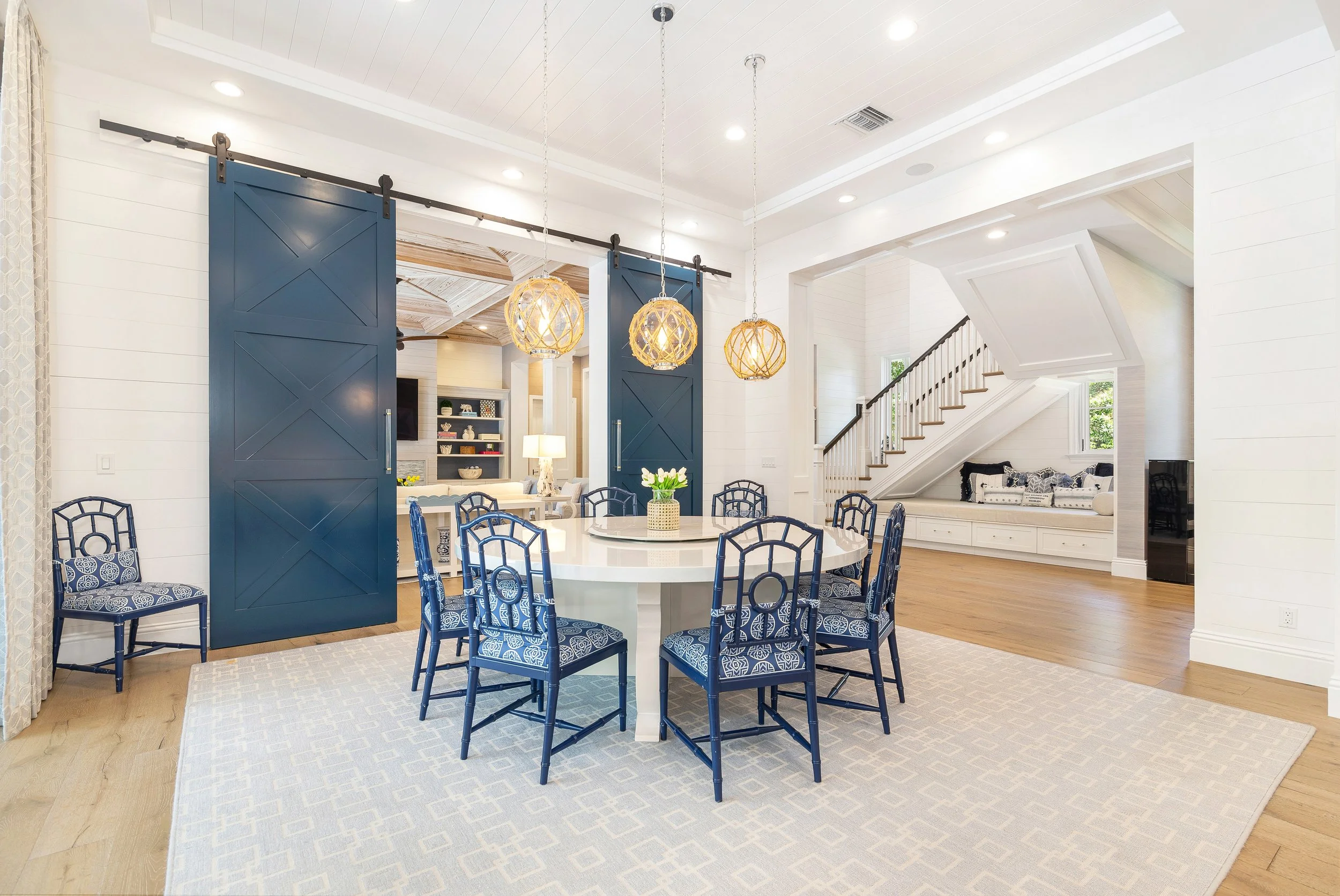 A spacious dining area with a white round table and blue patterned chairs on a light-colored rug. Blue sliding barn doors are hung from a black track, separating the dining area from a living space with a staircase. The ceiling has recessed lighting 