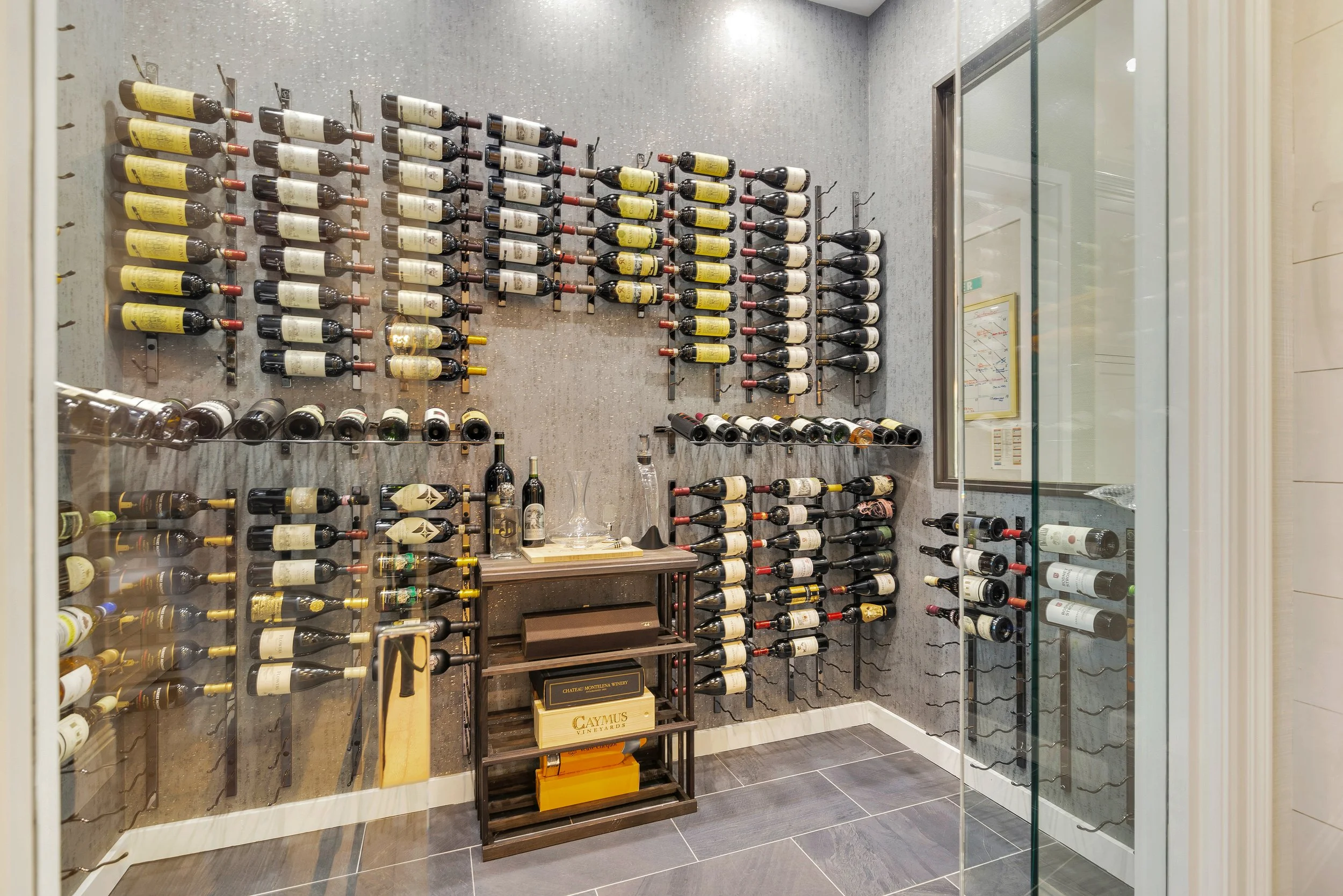 Display of wine bottles on wall-mounted racks inside a glass-walled wine cellar or wine shop, with additional bottles and boxes below.