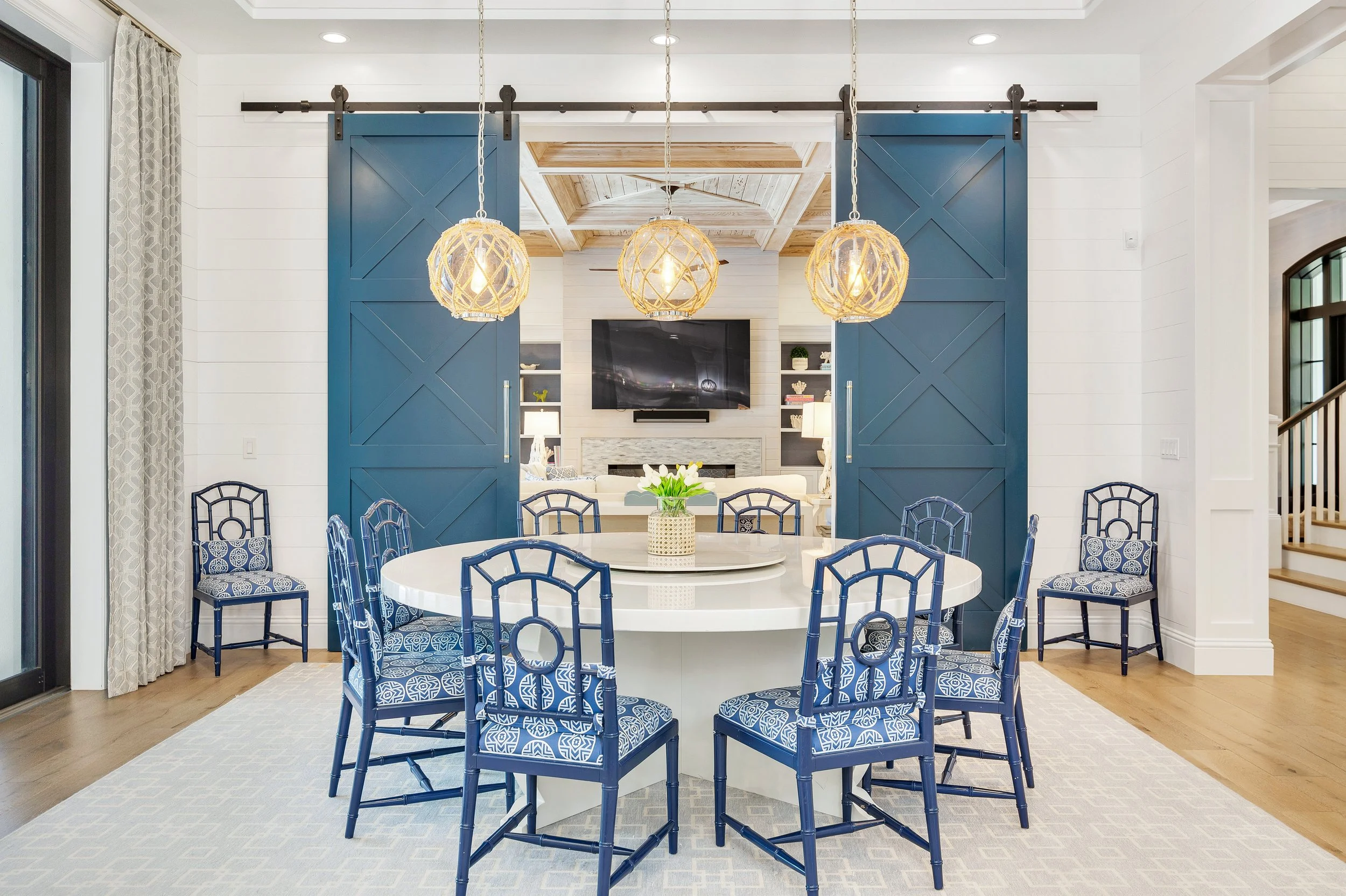 A dining room with a round white table surrounded by blue chairs with patterned cushions. Above the table are three woven pendant lights. In the background, there is a living room with a TV and shelves, visible through a pair of blue sliding barn doo
