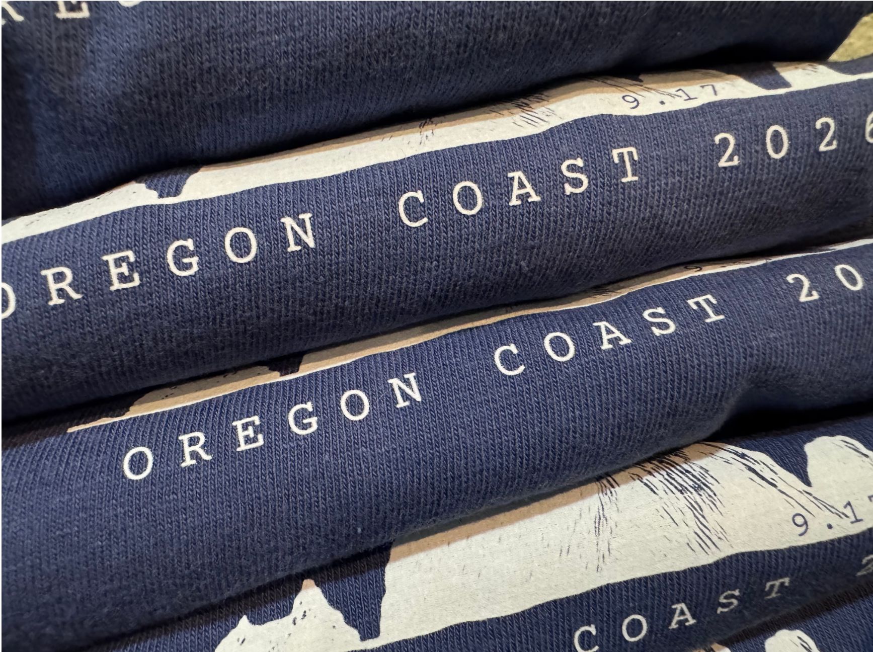 Stacked navy blue t-shirts with "Oregon Coast 2021" printed in white, laid on printed map paper.