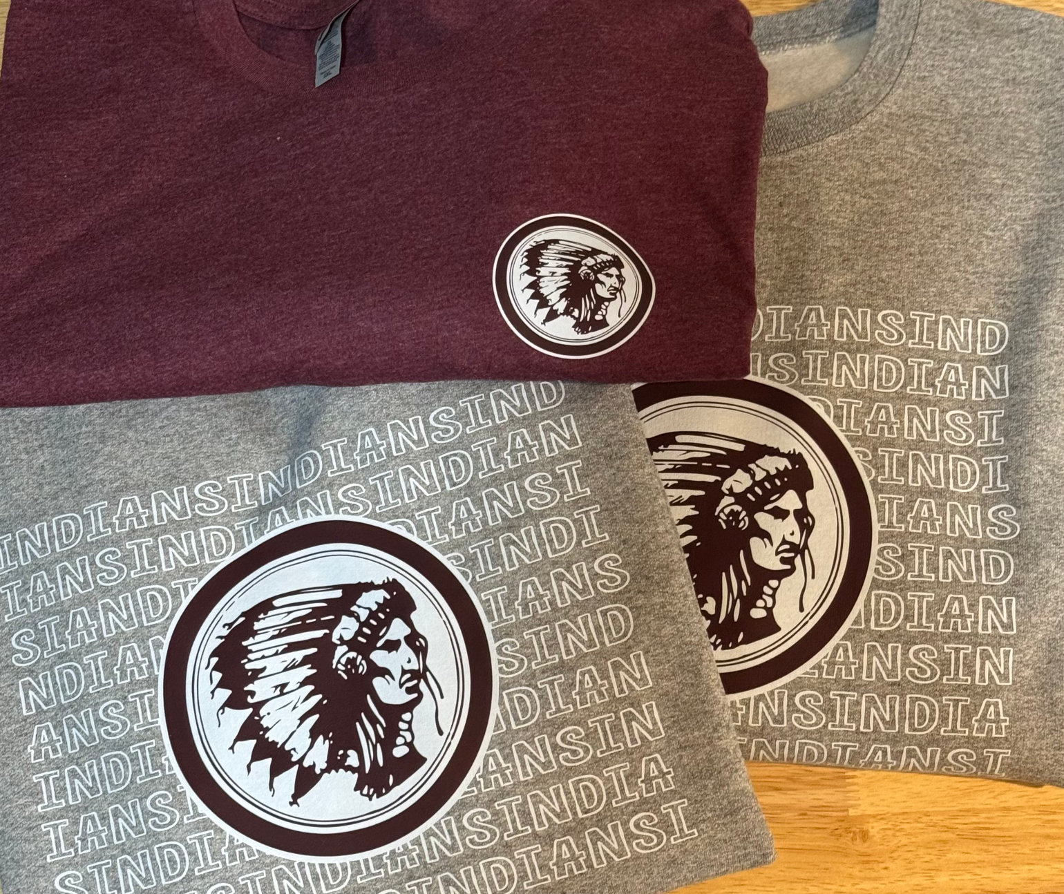 Two folded t-shirts, one dark red and one gray, featuring a logo of a Native American chief in profile with a feathered headdress, surrounded by repeated text saying 'INDIANA INDIAN'.
