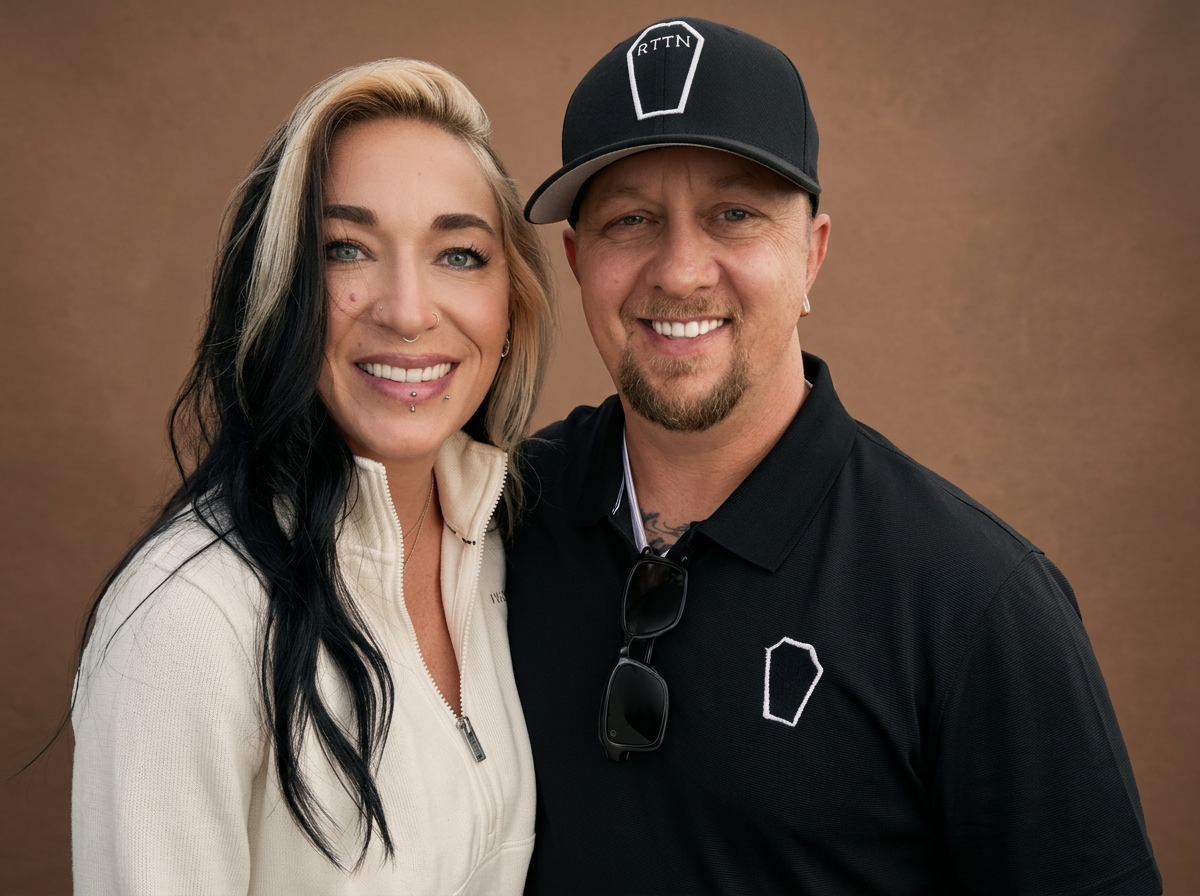 A smiling woman with blonde and black hair, wearing a cream-colored zip-up jacket, and a smiling man wearing a black cap and black polo shirt with sunglasses hanging from the shirt collar, standing close together against a plain background.