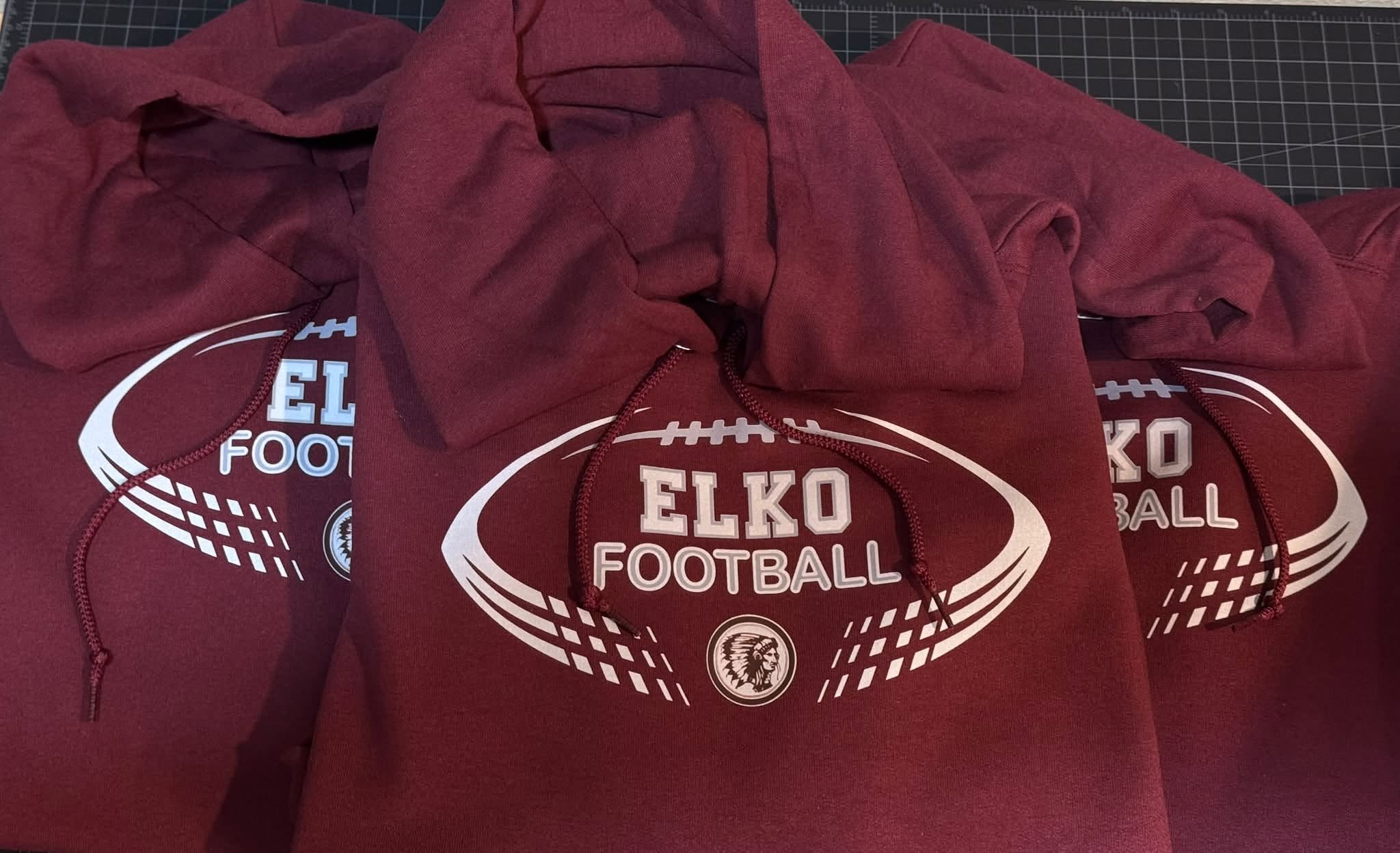 Red hoodie with a football graphic and the words 'Elko Football' on it, placed on top of other similar items with a cutting mat background.