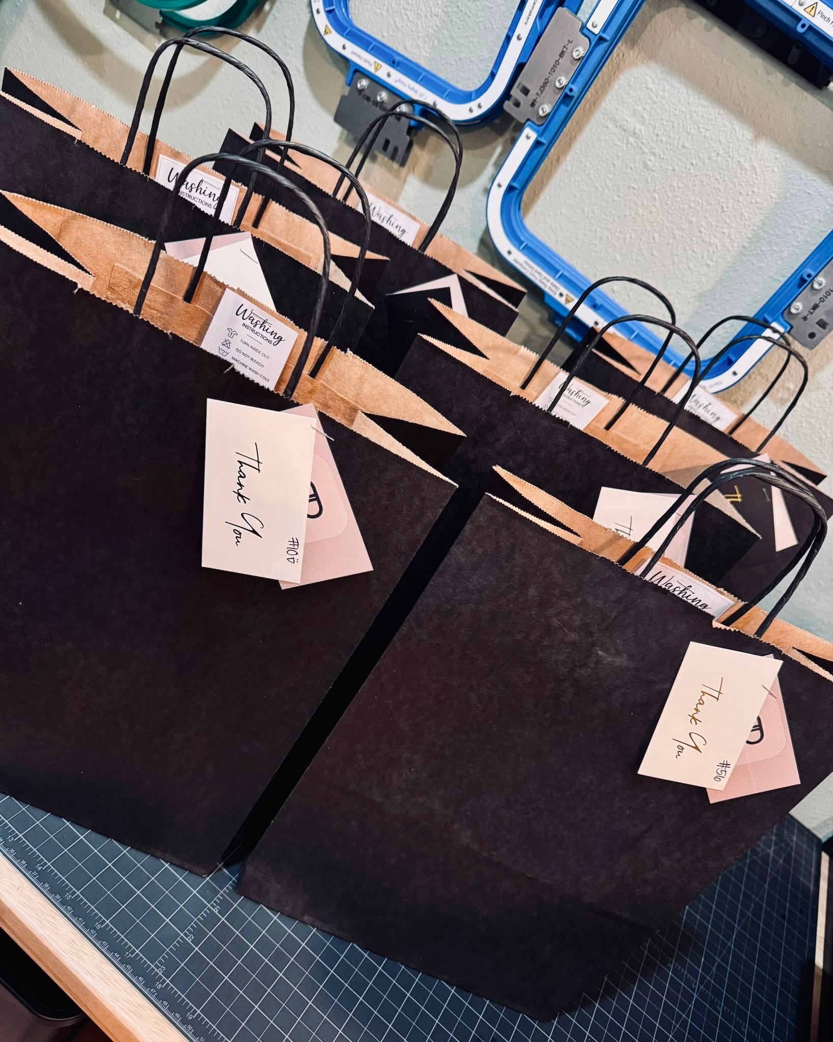 Multiple black and tan paper shopping bags with black handles and labels that read 'Washing Instructions' in a workspace on a table near a cutting mat. There are blue and black frame parts on the wall behind.