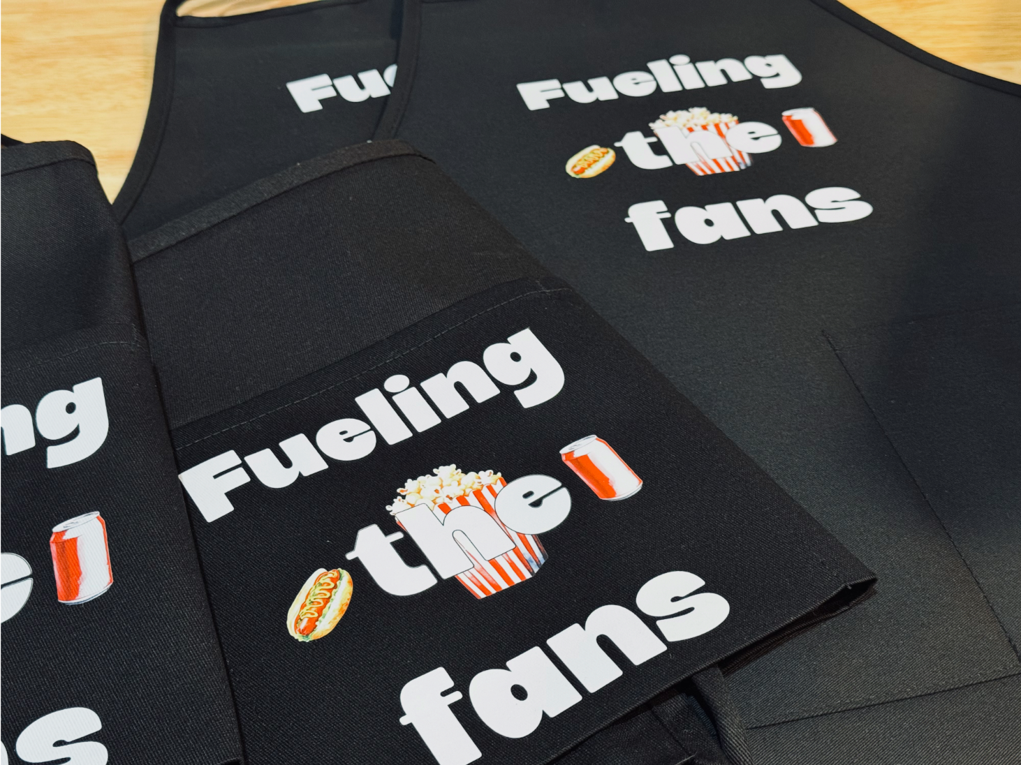 Black fabric item with white text that reads 'Fueling the fans', includes icons of a hot dog, popcorn, and a soda can, placed on a wooden surface.