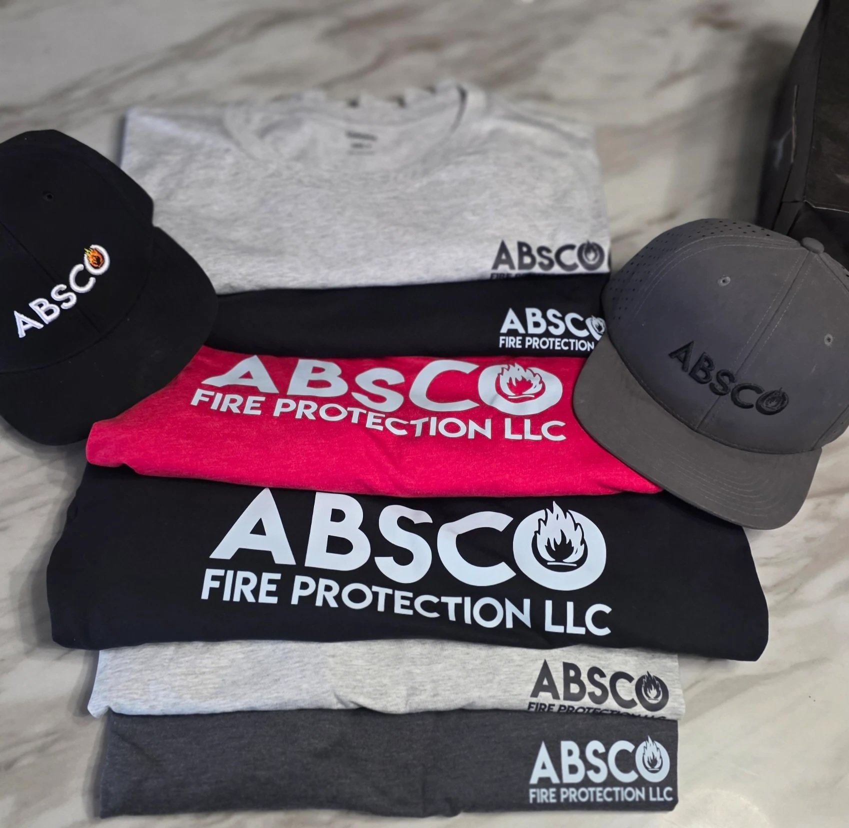 Various black, gray, red, and white T-shirts and caps with the logo and name "ABSC Fire Protection LLC" on them, displayed on a surface.