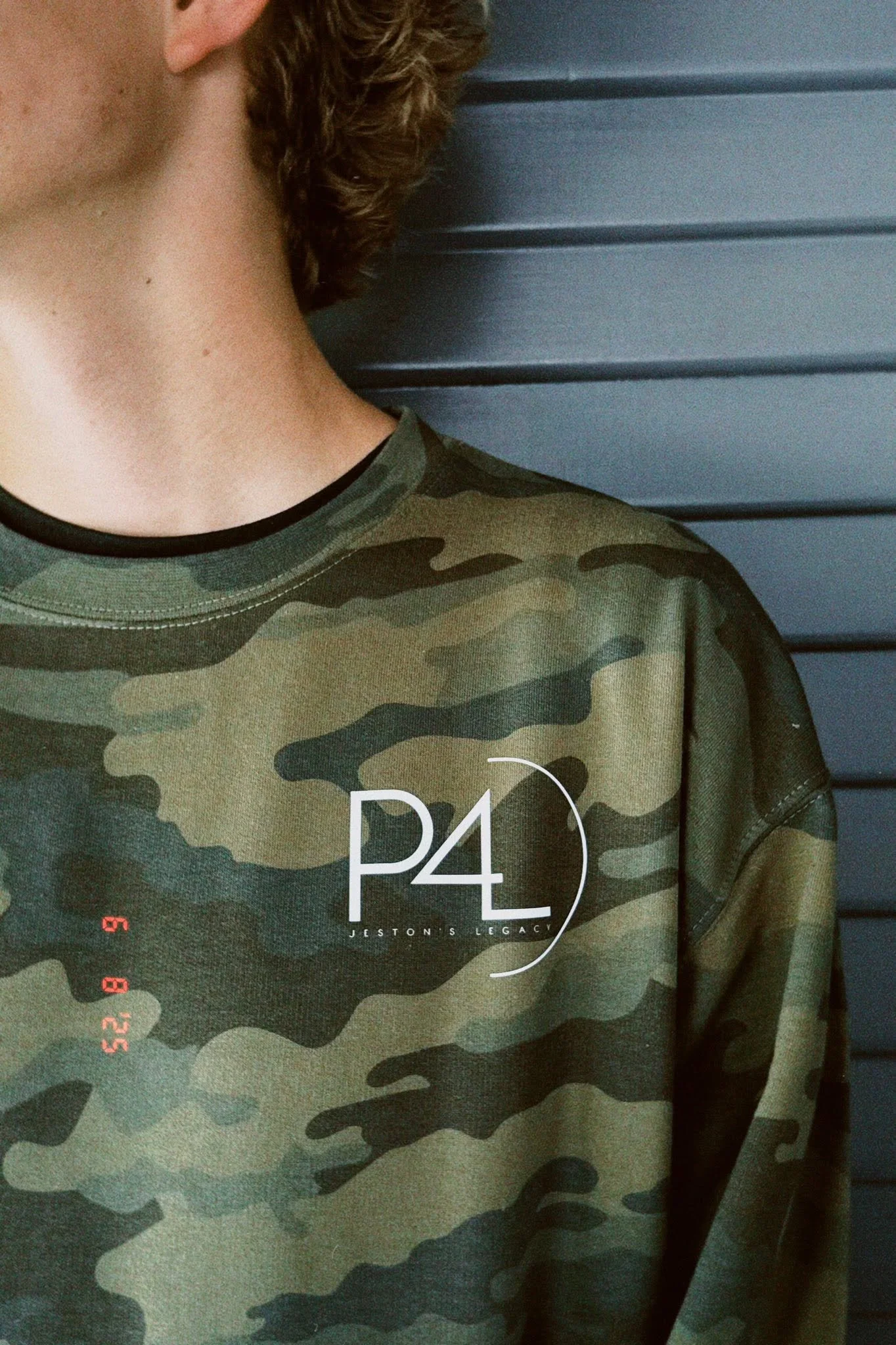 Close-up of a person wearing a camouflage shirt with the text 'P4L' and 'JESTON'S LEGACY' printed on it. The person's face is not visible, and they are standing in front of a dark gray horizontal panel.