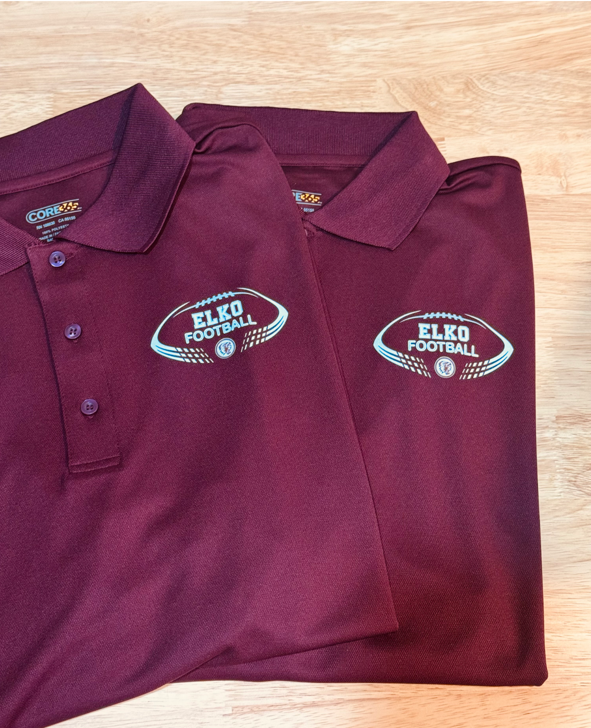 Two maroon polo shirts with 'Elko Football' and a football graphic logo embroidered on them, placed on a light wooden surface.
