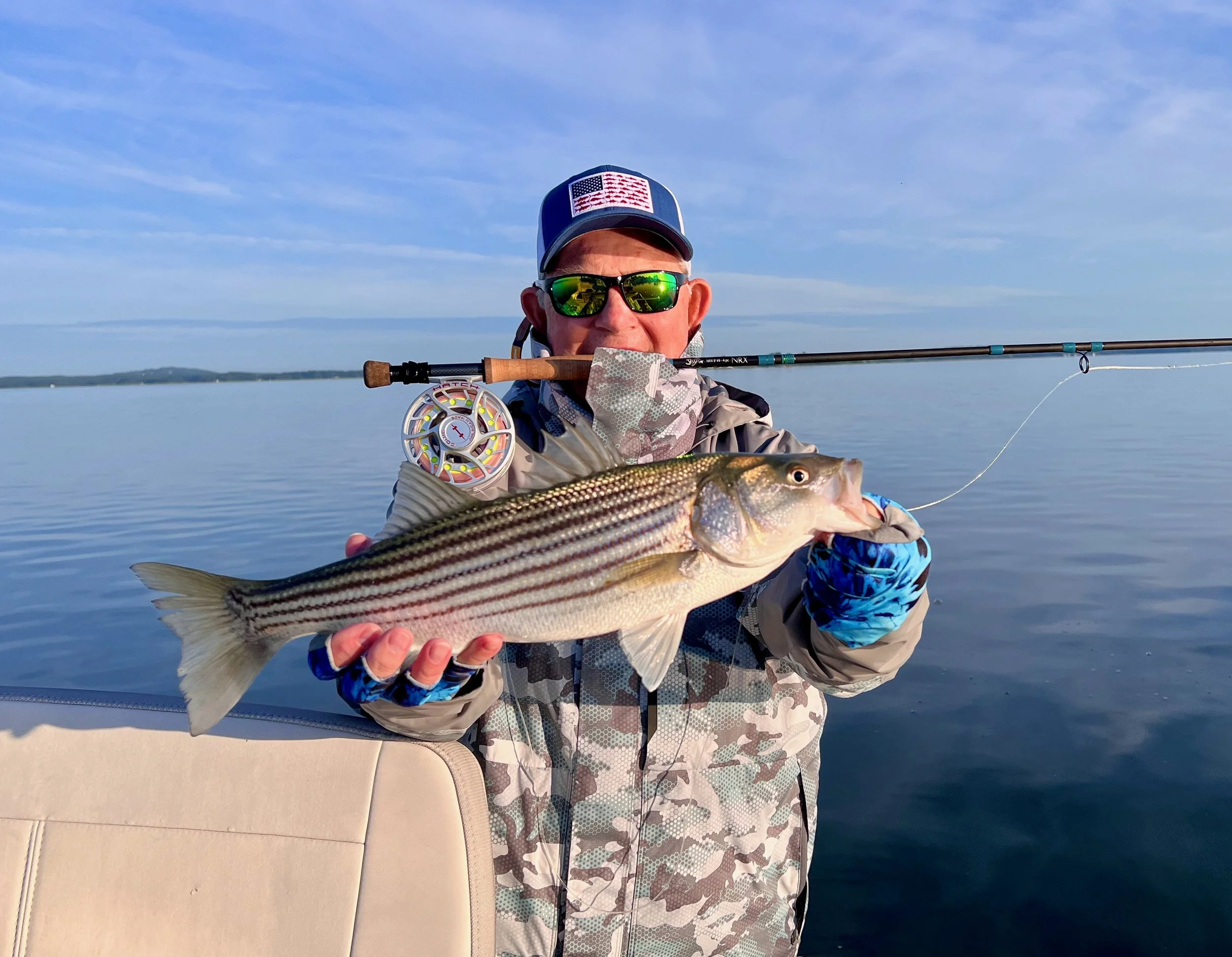 A person fishing on a boat, holding a large fish over the side. The person is wearing sunglasses, a camouflage jacket, and a hat with an American flag patch. The water and sky are visible in the background.