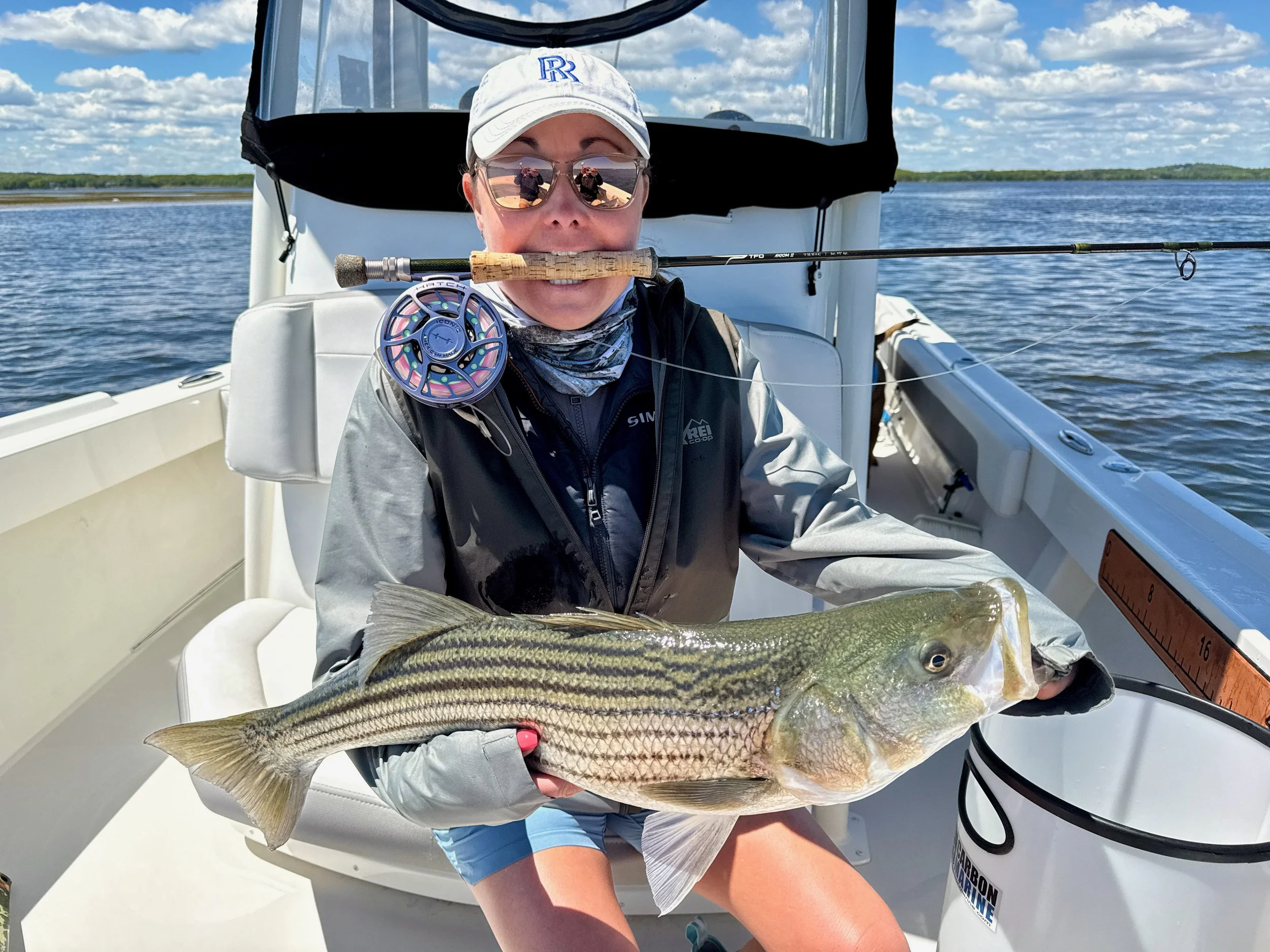 A woman on a boat holding a large fish she caught with a fishing rod in her mouth, wearing sunglasses, a white cap, and a gray jacket, with a blue sky and water in the background.
