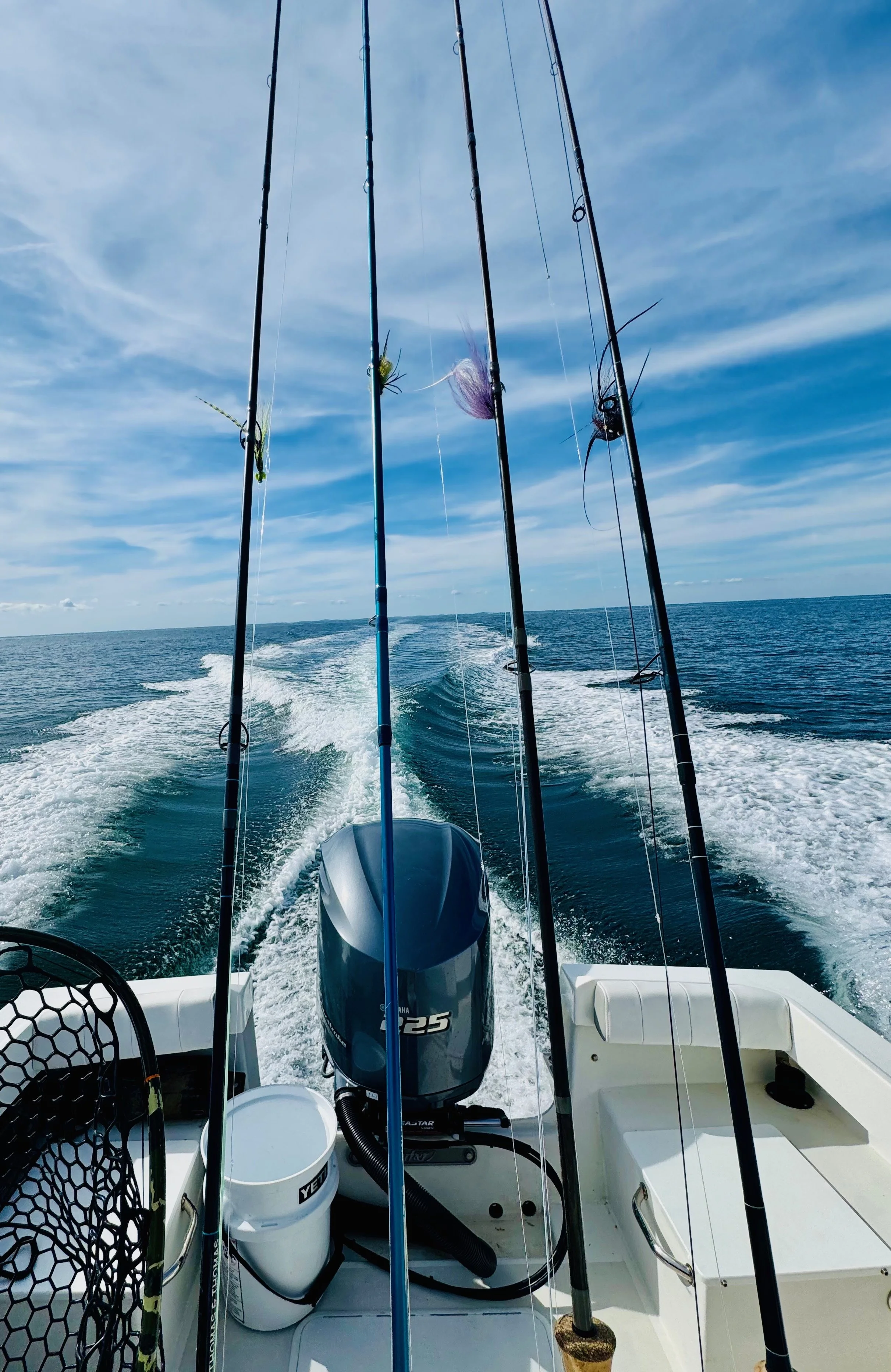 View from the back of a boat with several fishing rods mounted, water in motion behind the boat, and a partly cloudy sky overhead.