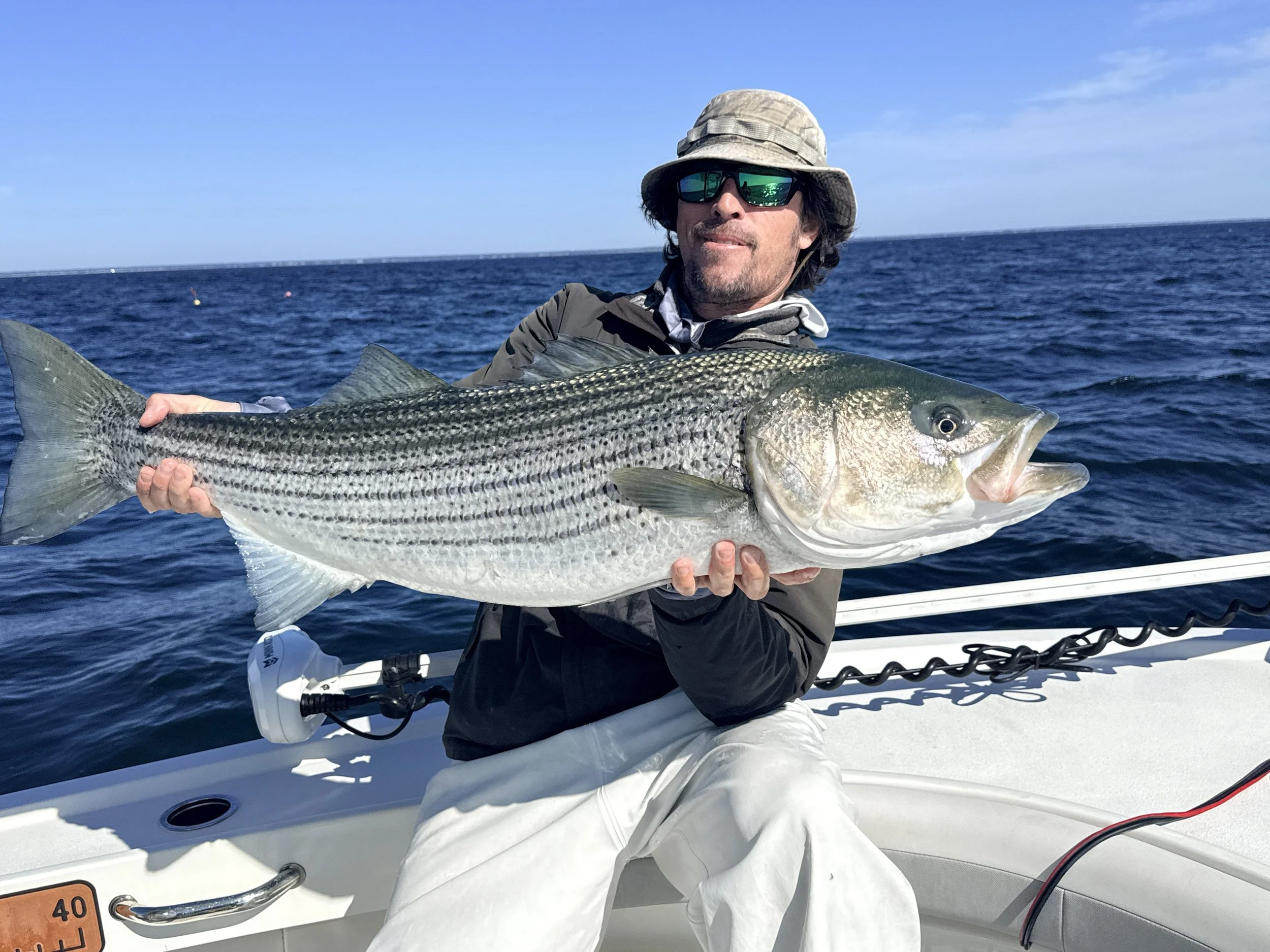 A man wearing sunglasses, a hat, and a jacket is holding a large fish on a boat in the open sea, with a clear blue sky in the background.