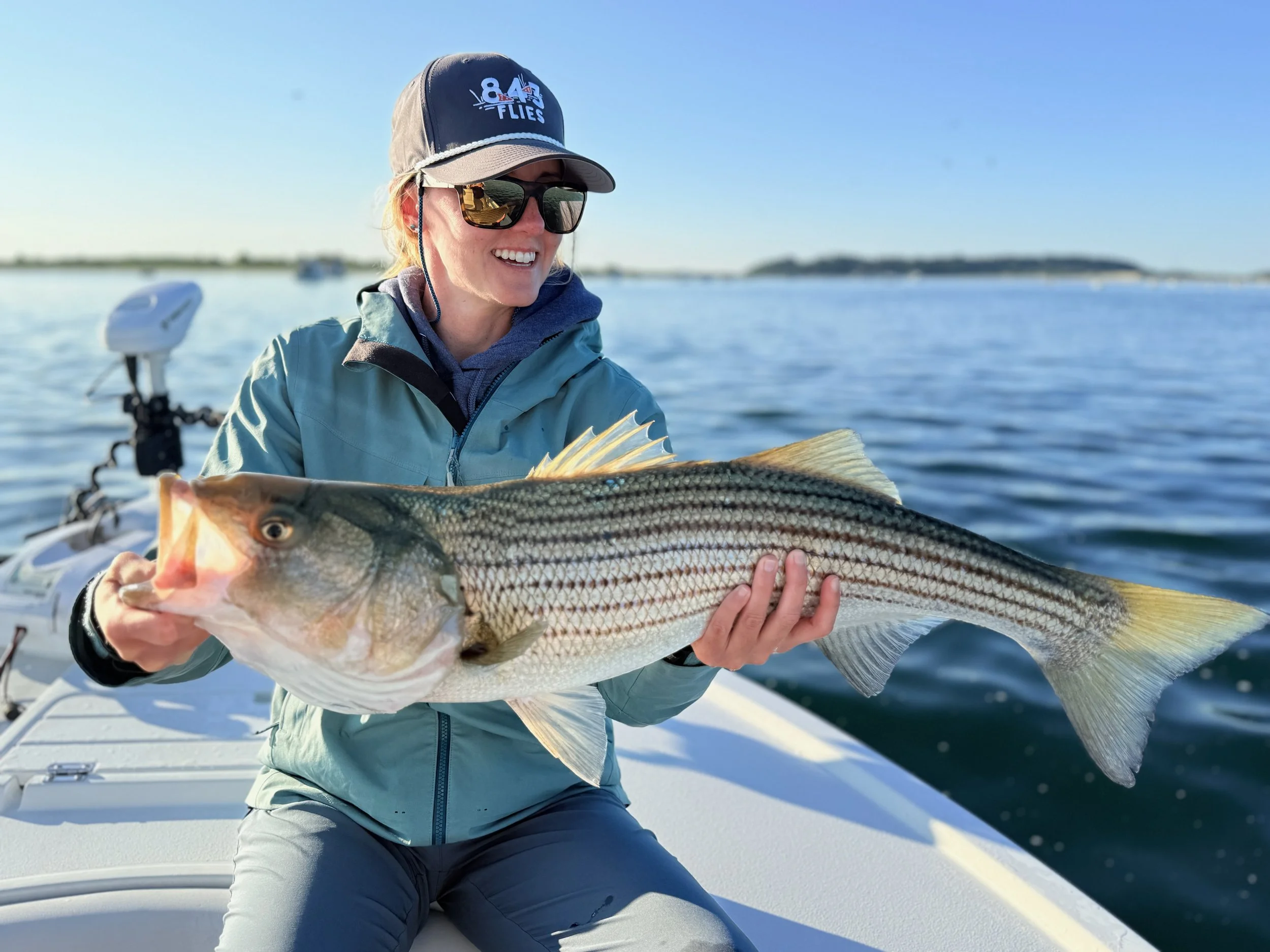 A woman on a boat holding a large fish she caught, smiling and wearing sunglasses, a cap, and outdoor clothing with a body of water and distant land in the background.
