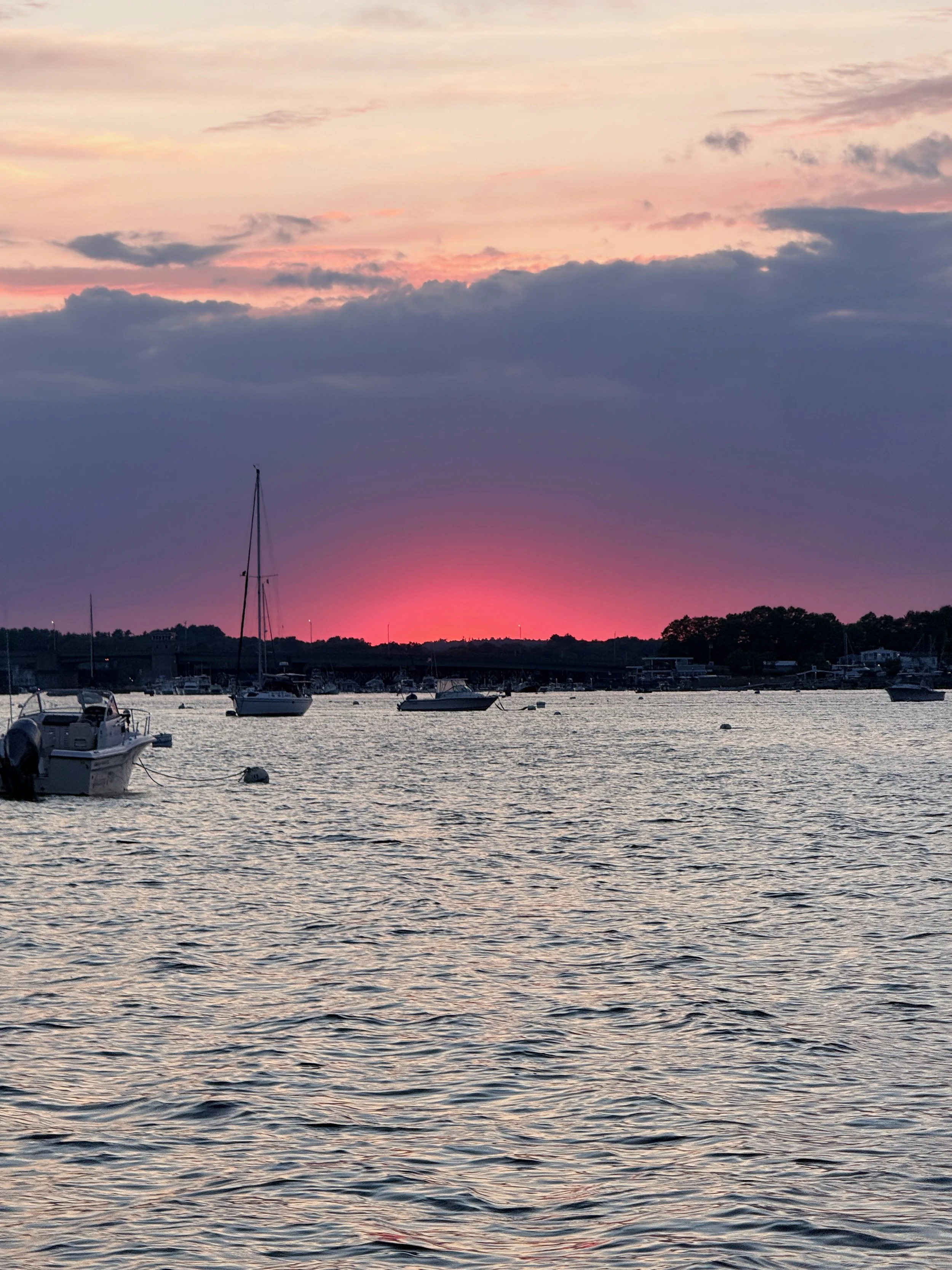 A tranquil harbor scene at sunset with boats anchored on calm water, and a colorful sky with pink, orange, and purple clouds.