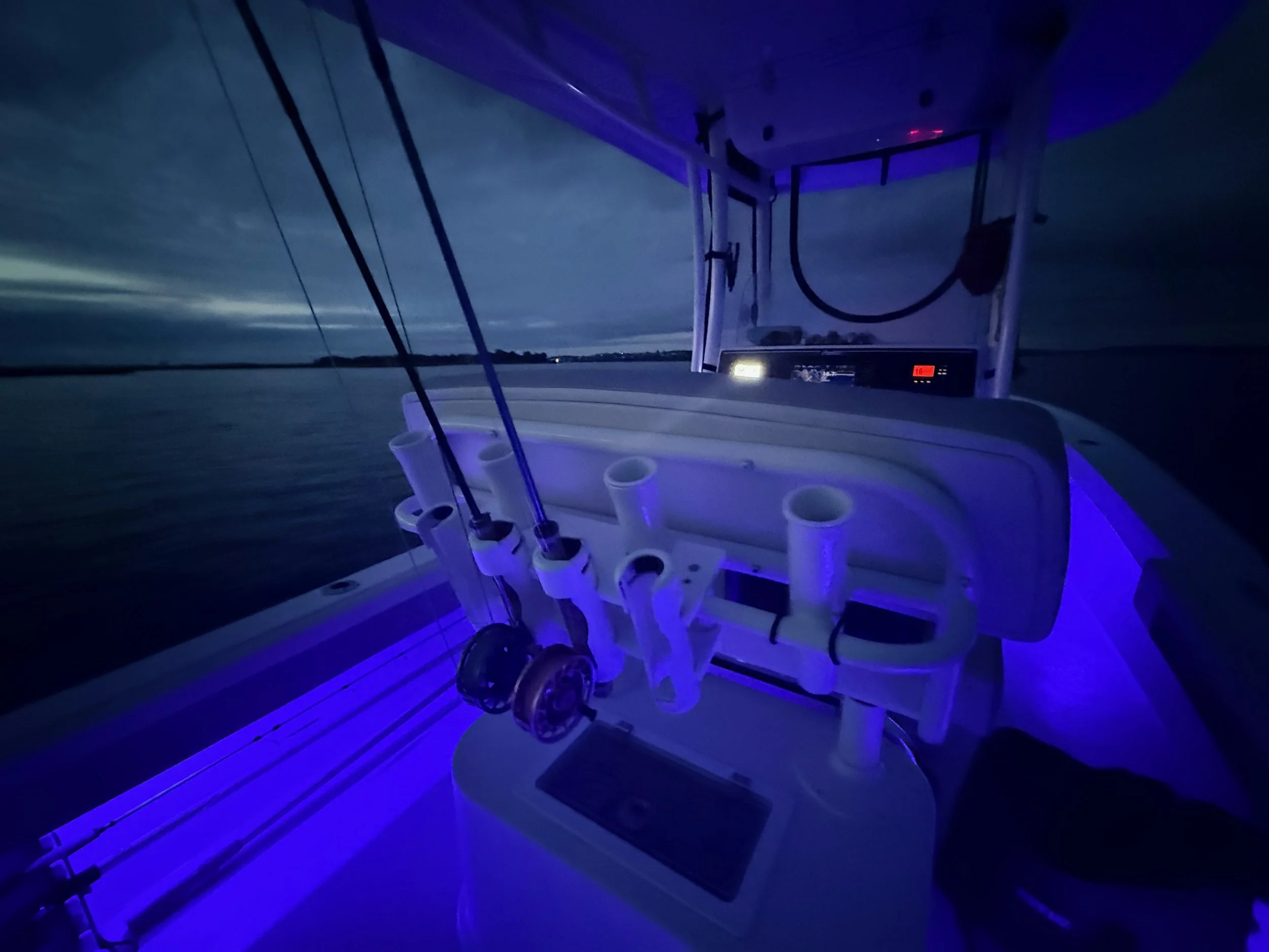 Nighttime scene from a boat with purple and blue lighting, showing fishing rods and a water body in the background.