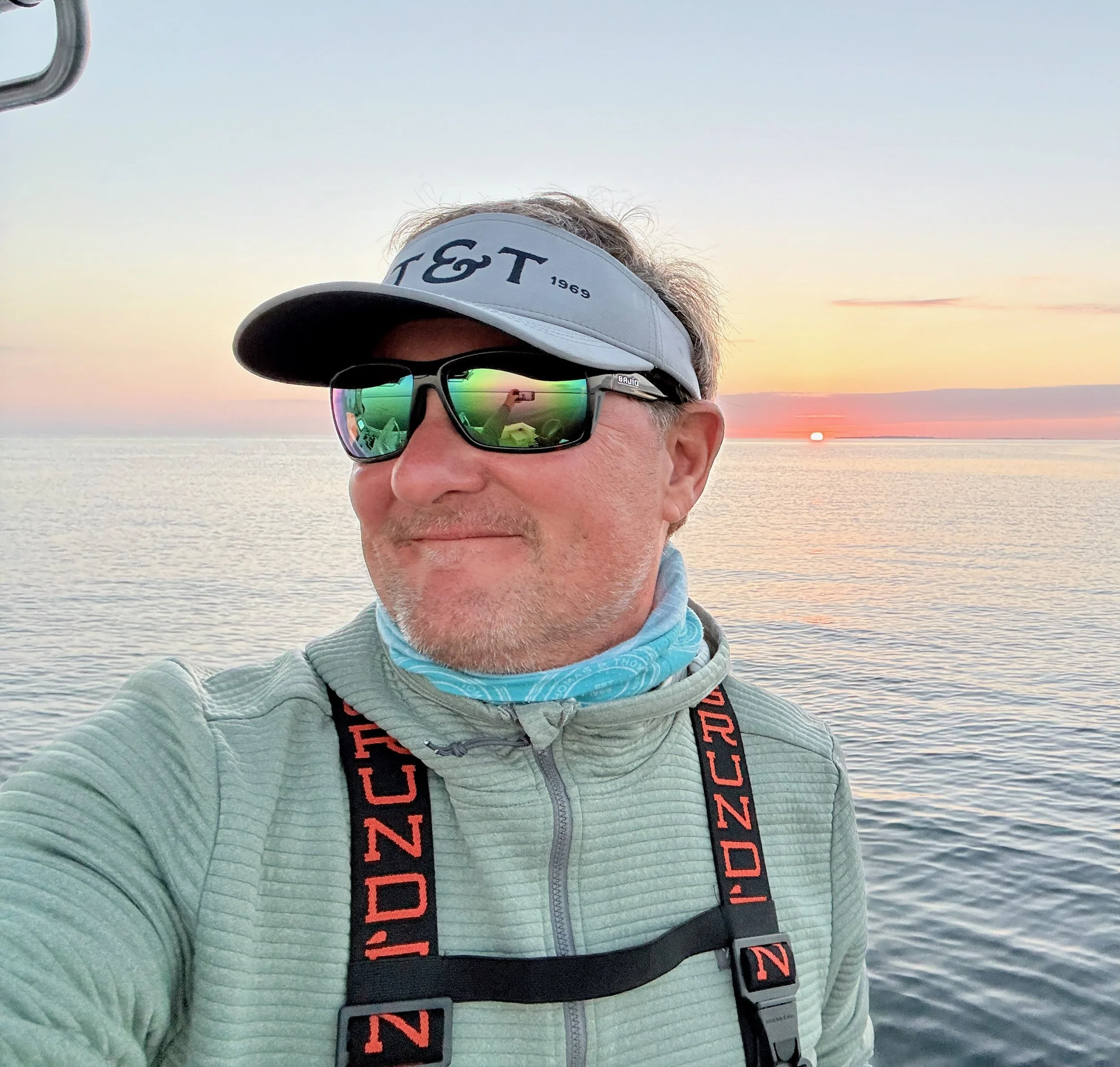 A man wearing a white visor, reflective sunglasses, and a light gray jacket takes a selfie in front of a sunset over water. The sunrise or sunset is low on the horizon with pink and orange hues in the sky.