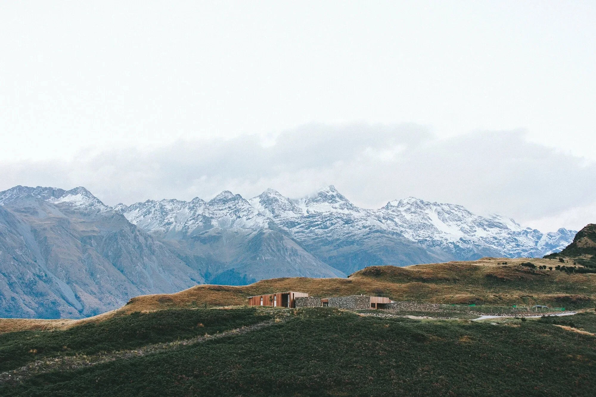 A mountainous landscape with snow-capped peaks, rolling green hills in the foreground, and a modern building at the base of the hills. Overcast sky with clouds covering the mountain peaks.