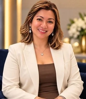 A woman with shoulder-length hair, wearing a beige blazer and a brown top, sitting and smiling in an indoor setting with a blurred background of flowers and decor.