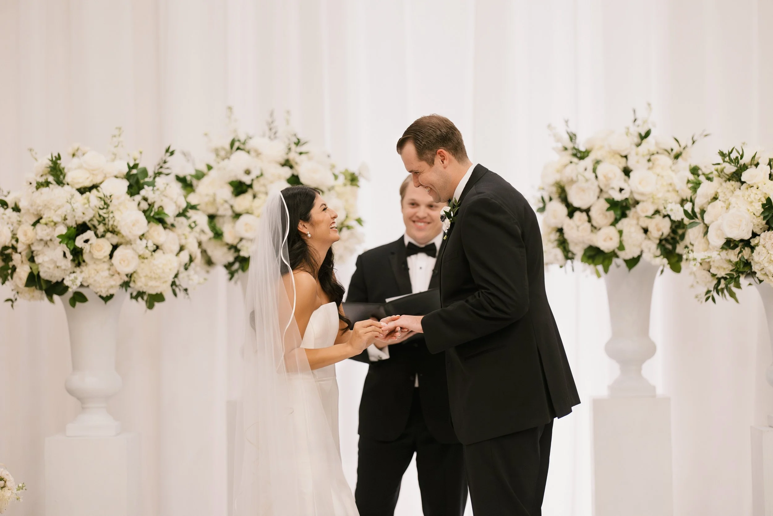 A bride and groom holding hands during their wedding ceremony, smiling at each other, with an officiant behind them, surrounded by large white floral arrangements.