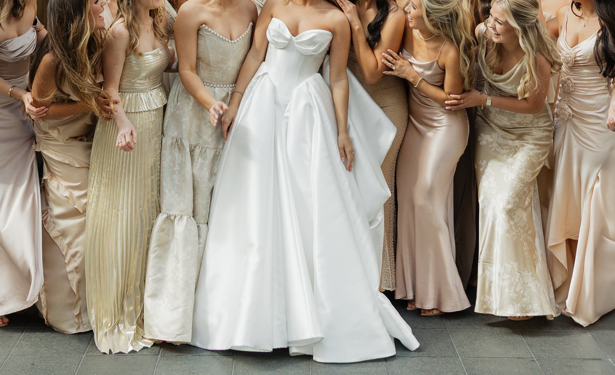 Group of women in satin and lace dresses gathered together at a wedding, centering on the bride in a strapless white gown.