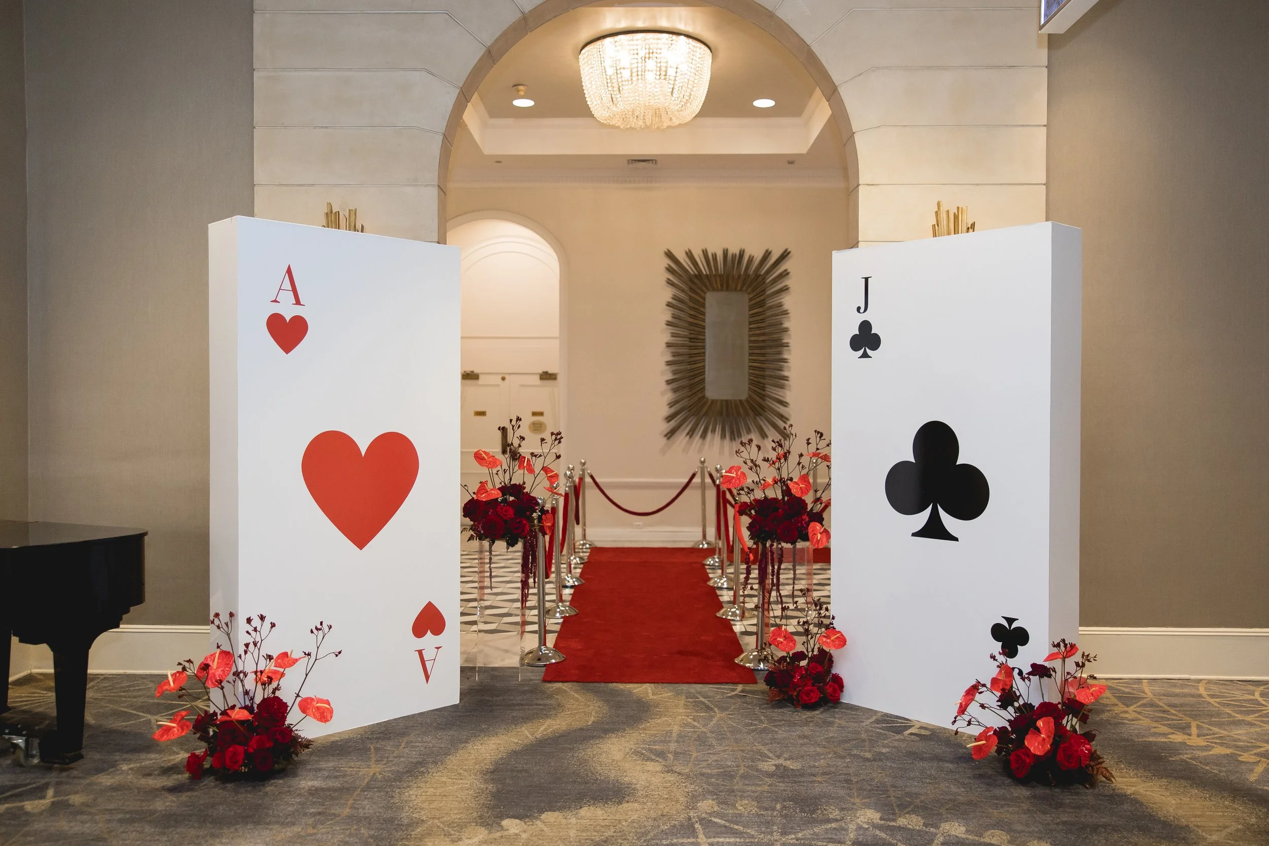 Entrance decorated with oversized playing cards, the Ace of Hearts and Jack of Clubs, flanked by red flowers and an aisle with a red carpet leading to a mirror and elegant chandelier.