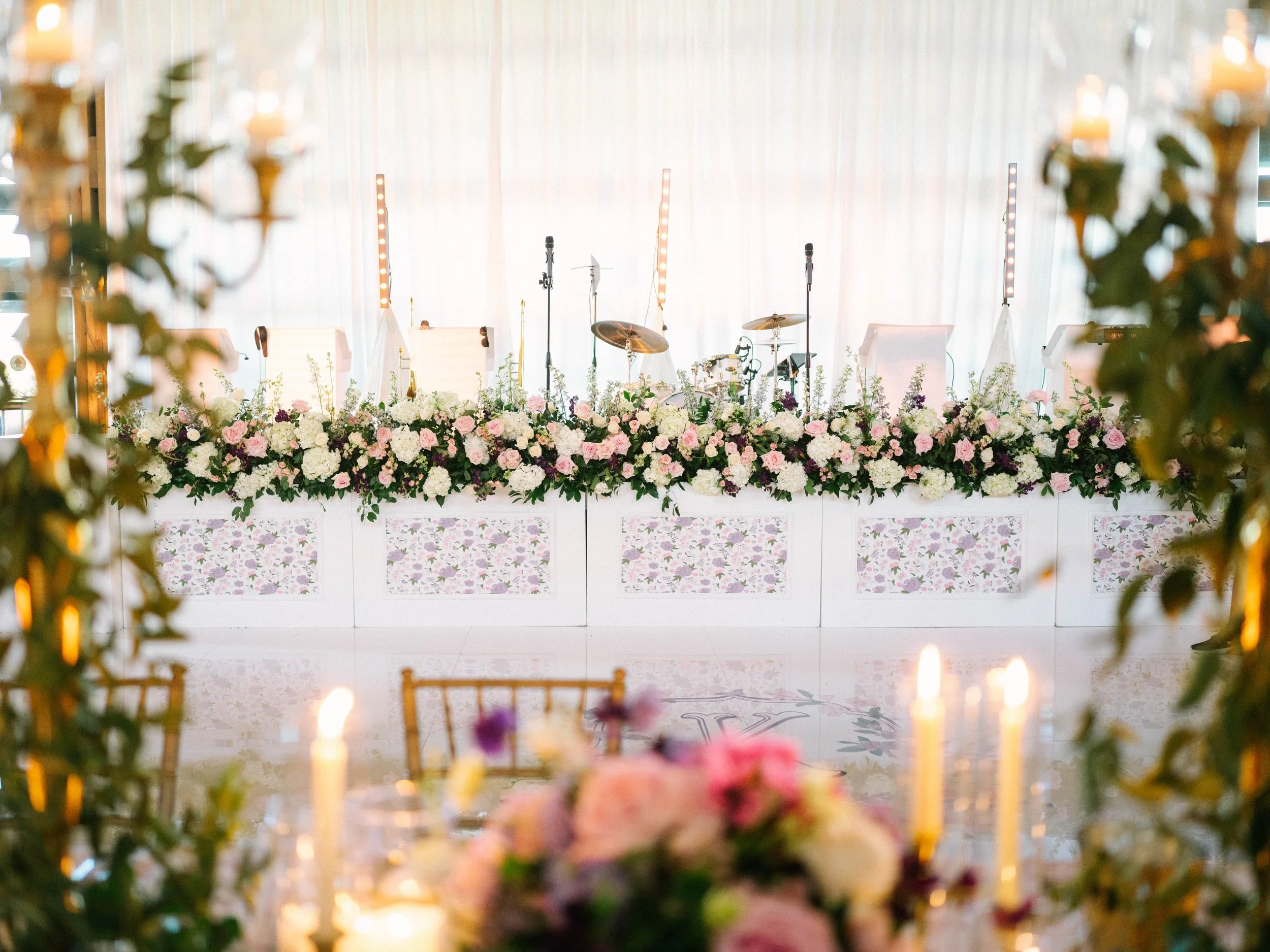 Wedding or event stage decorated with white and pink flowers, with four chairs, music stands, microphones, and musical instruments in the background, with blurred flowers and candles in the foreground.