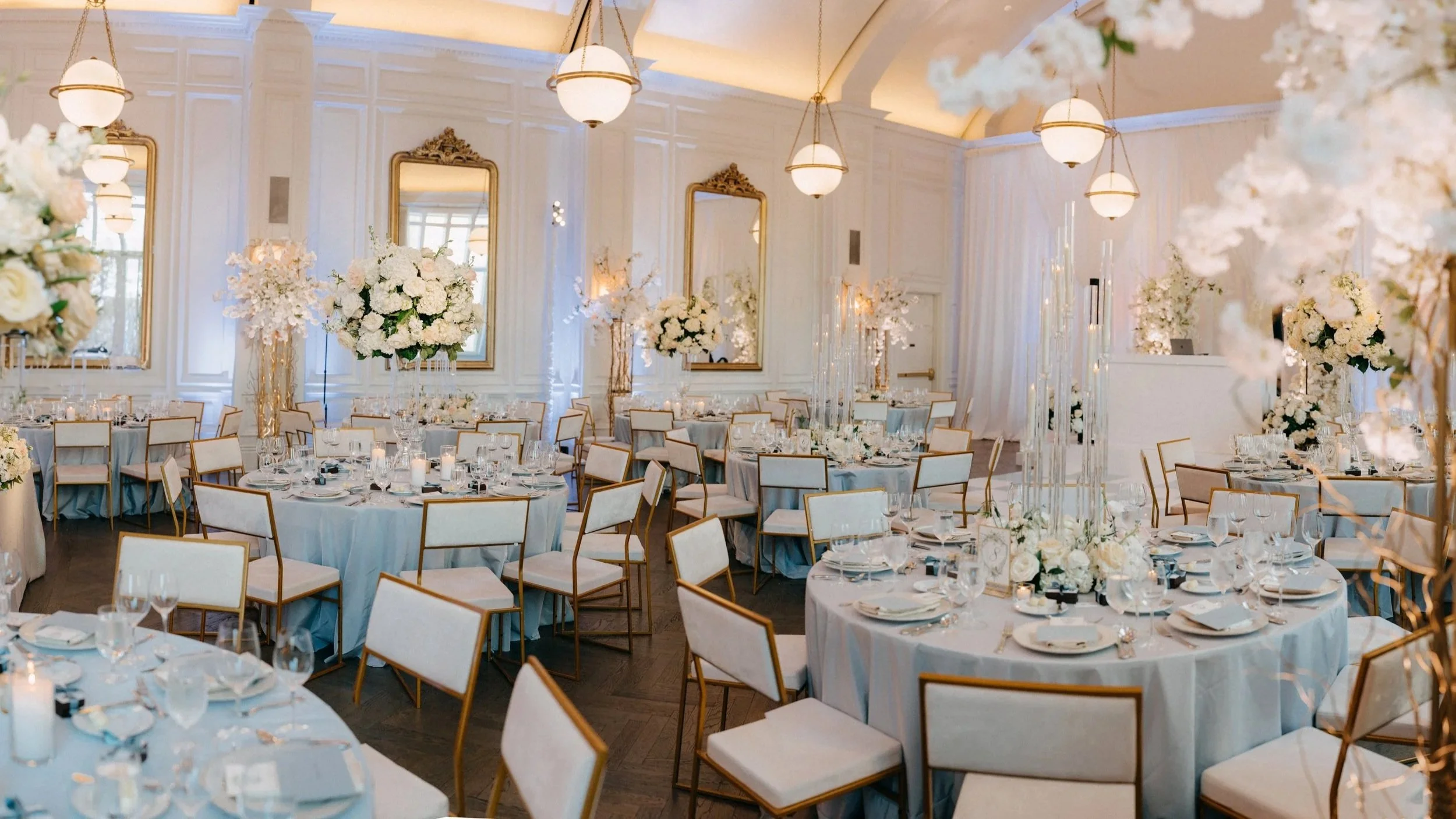 Elegant banquet hall decorated for a wedding or formal event with white and cream floral centerpieces, gold accents on chairs and mirrors, round tables set with fine china and glassware, and soft lighting.