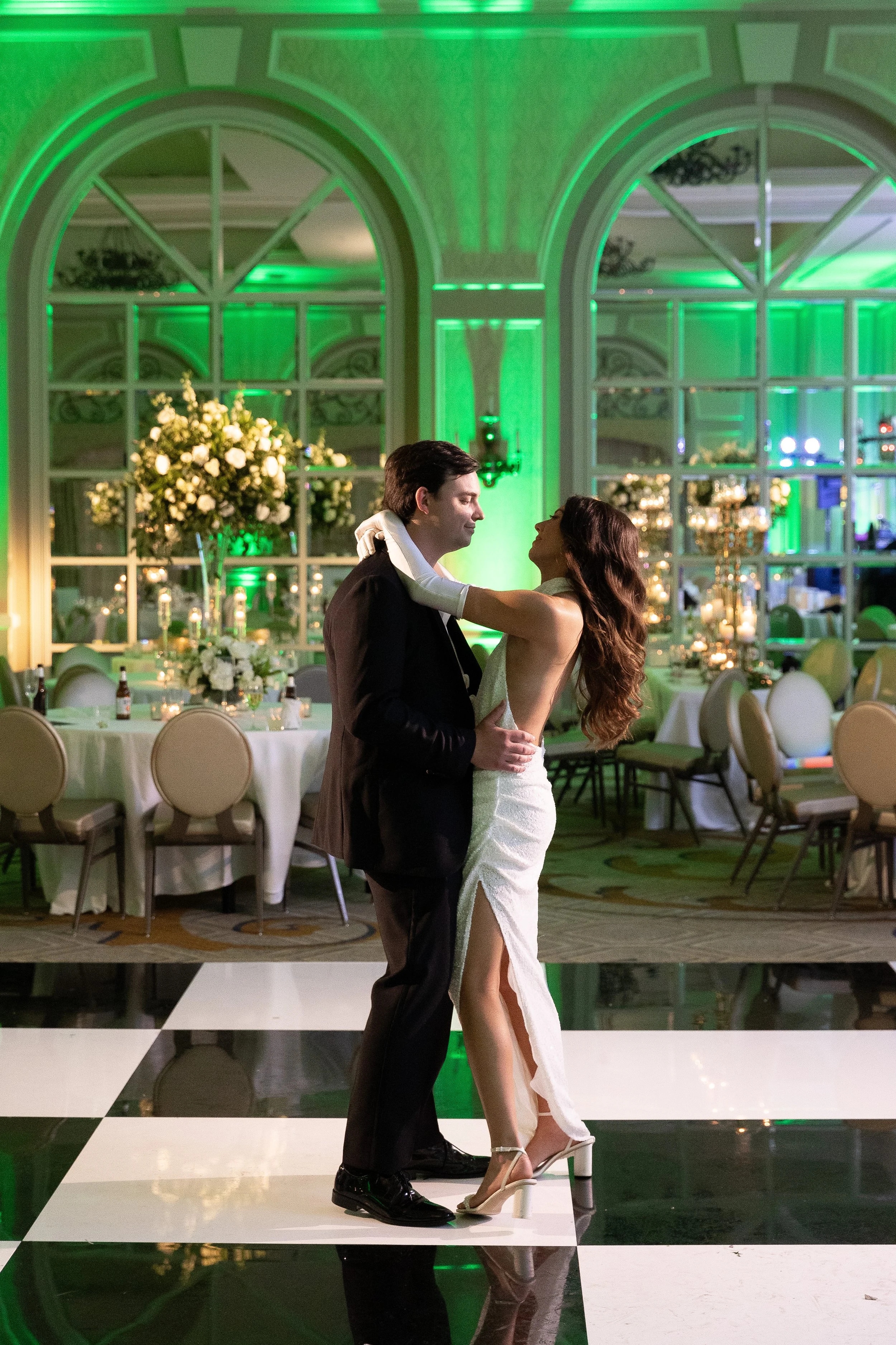 A man and woman dancing closely at a wedding reception in an elegant, decorated ballroom with floral centerpieces and green ambient lighting.