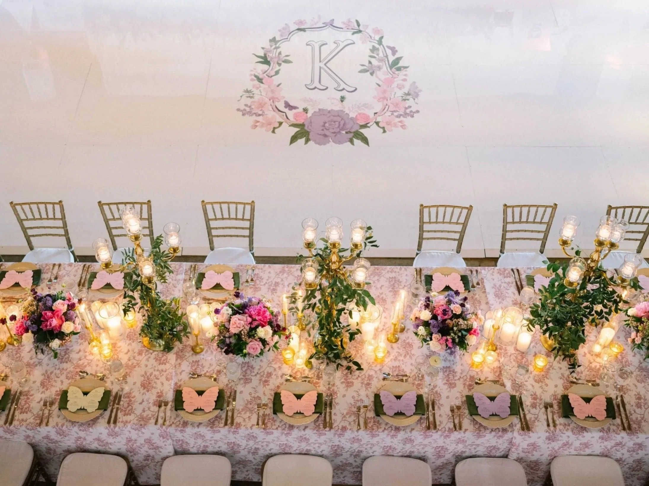 A decorated banquet table with pink and purple floral centerpieces, gold candlesticks, and butterfly-shaped place cards, set for an event or celebration, viewed from above.