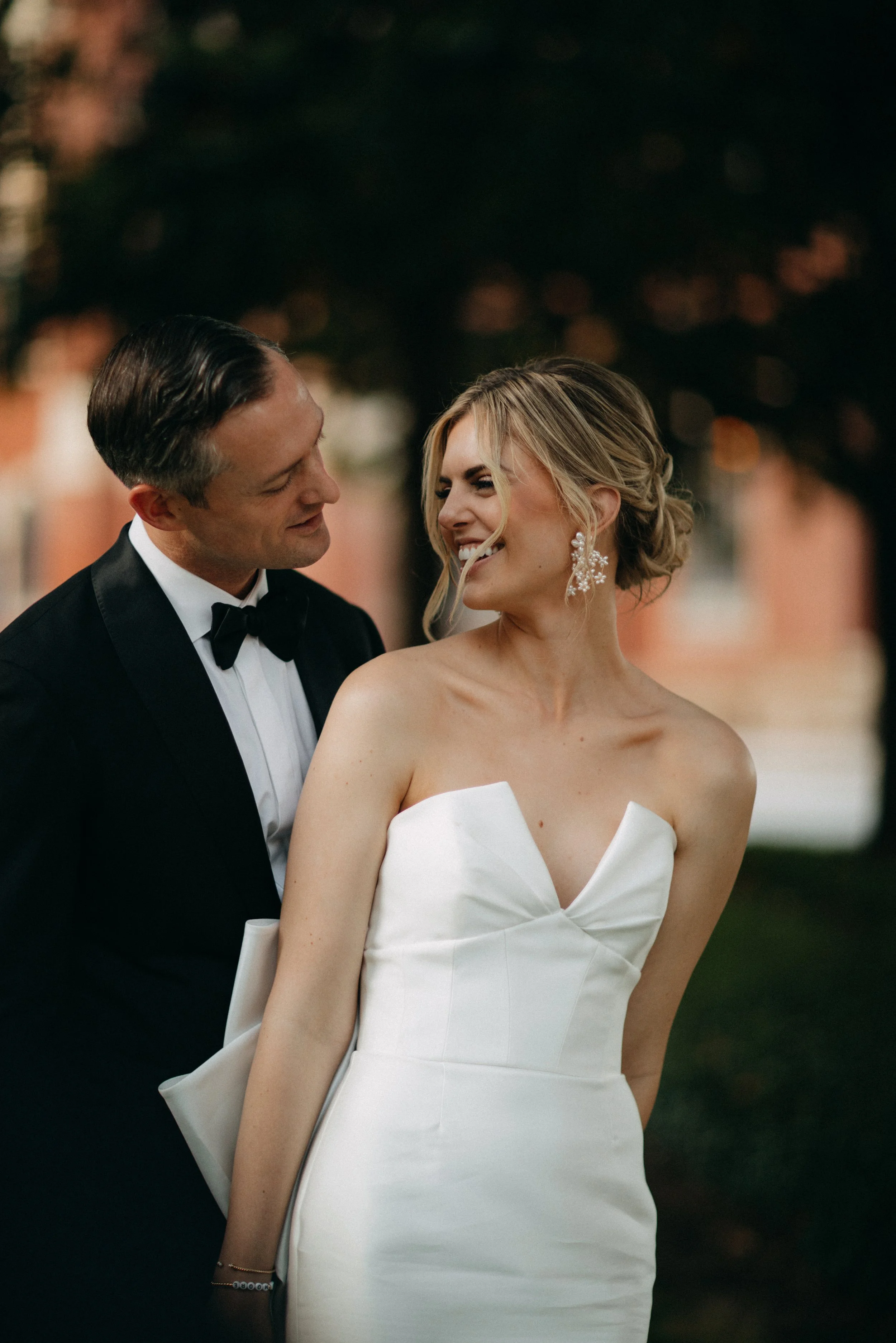A bride and groom smiling and looking at each other outdoors during sunset, with the bride wearing a strapless white wedding gown and the groom in a black tuxedo.