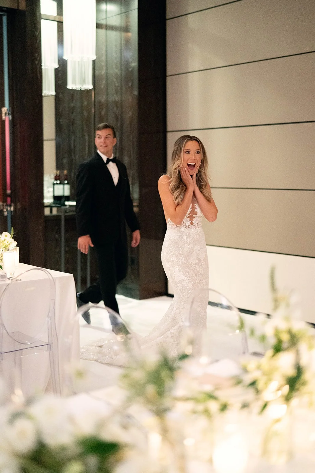 A bride in a white lace wedding dress looks surprised and happy, standing in a reception hall with a groom in a tuxedo in the background.