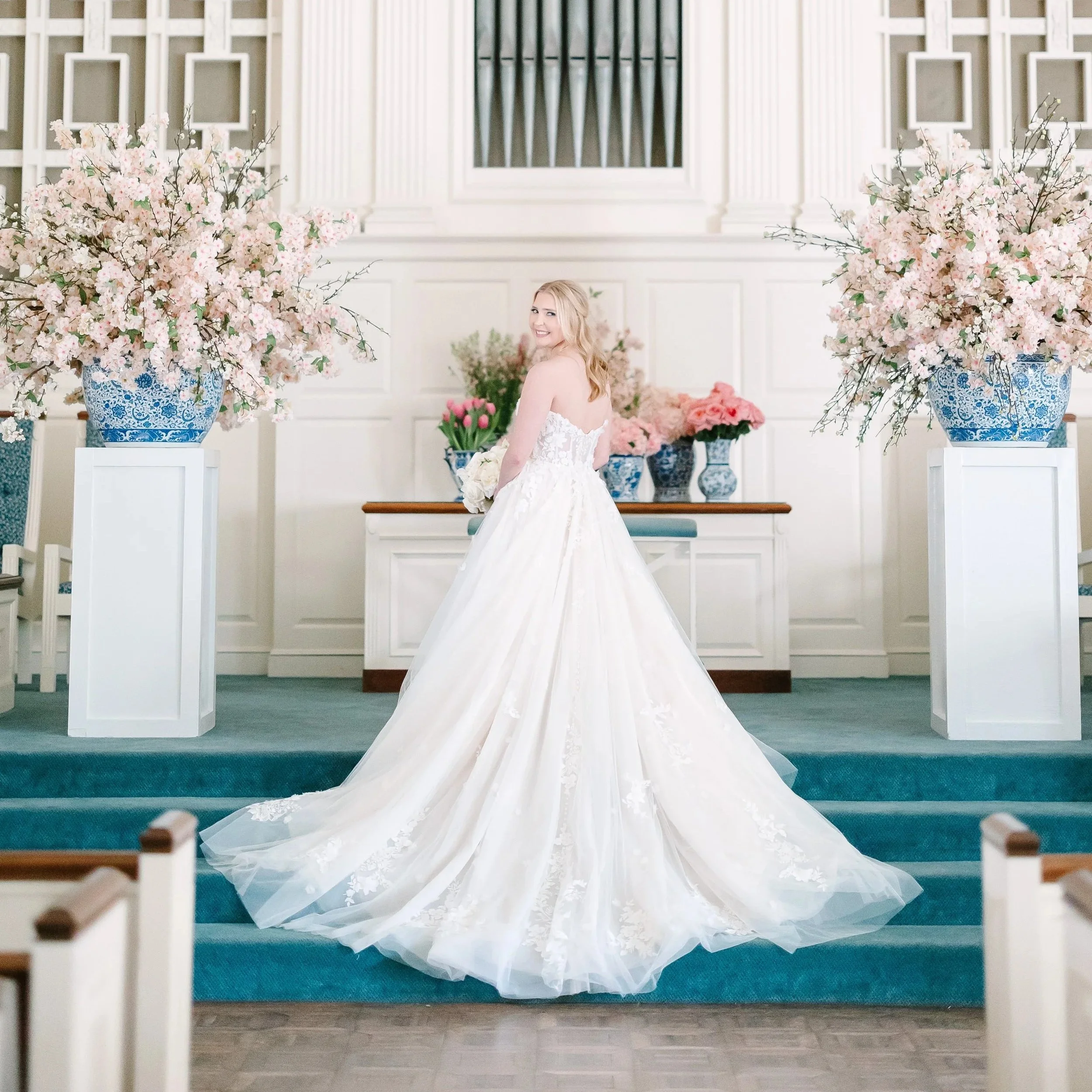 A bride in a white wedding gown standing on steps inside a decorated wedding chapel, holding a bouquet and smiling.