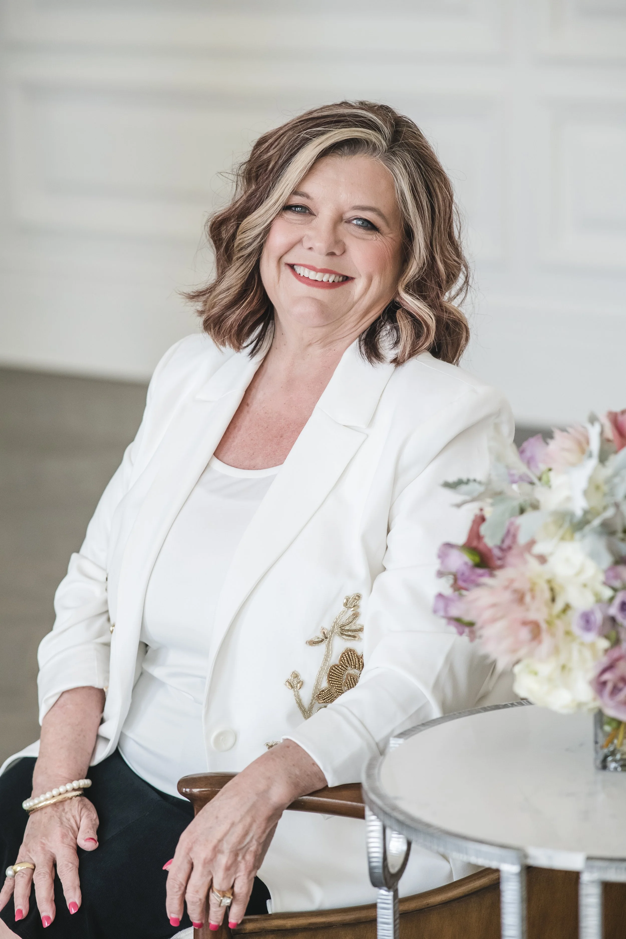 A woman with shoulder-length wavy brown hair with blonde highlights, smiling while sitting on a chair. She is dressed in a white blazer with embroidery, white top, and black pants. There is a bouquet of pink, white, and purple flowers on a small table beside her.