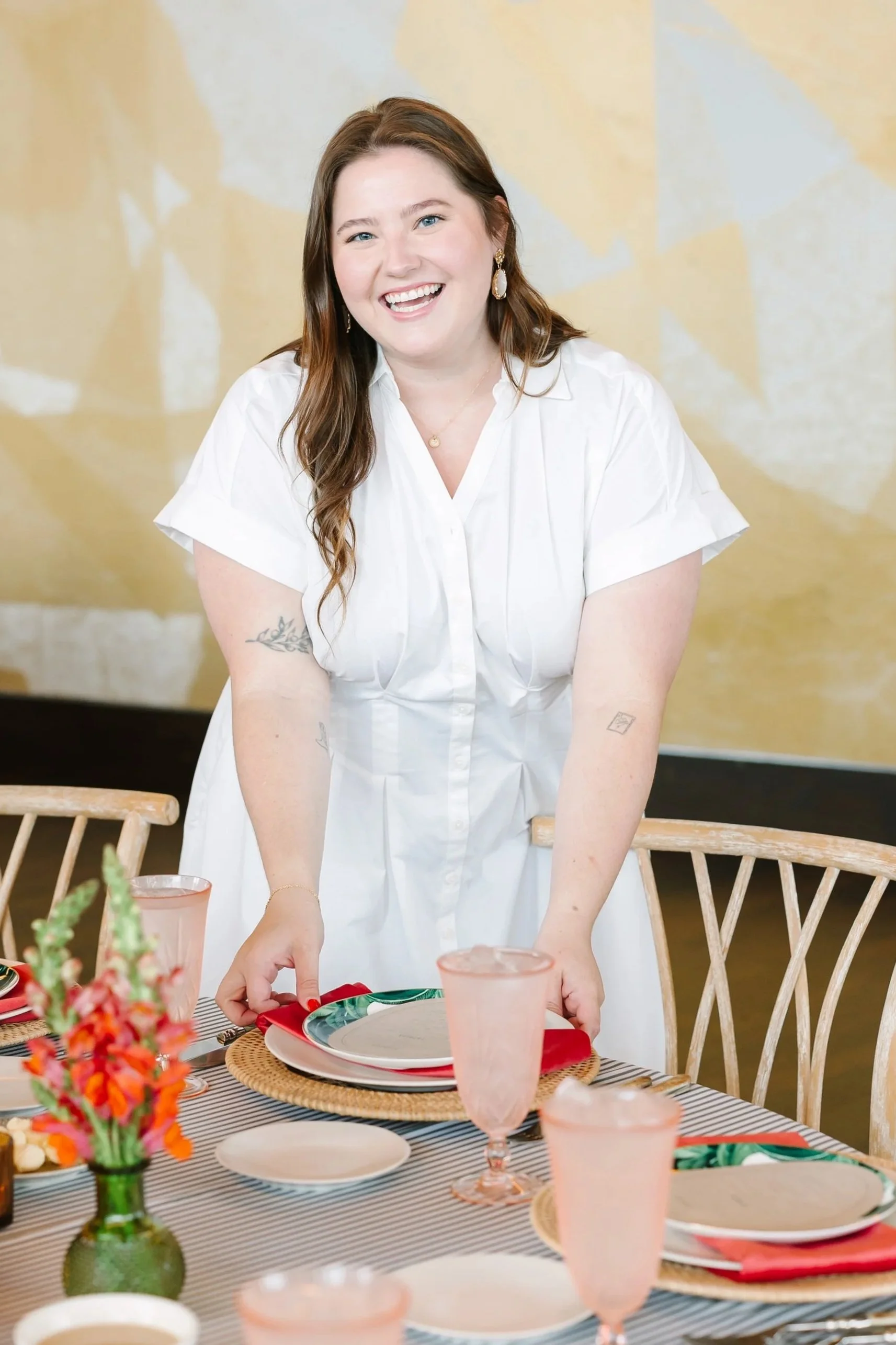 A smiling woman with long brown hair, wearing a white dress, standing behind a dining table set with plates, glasses, and a flower arrangement.