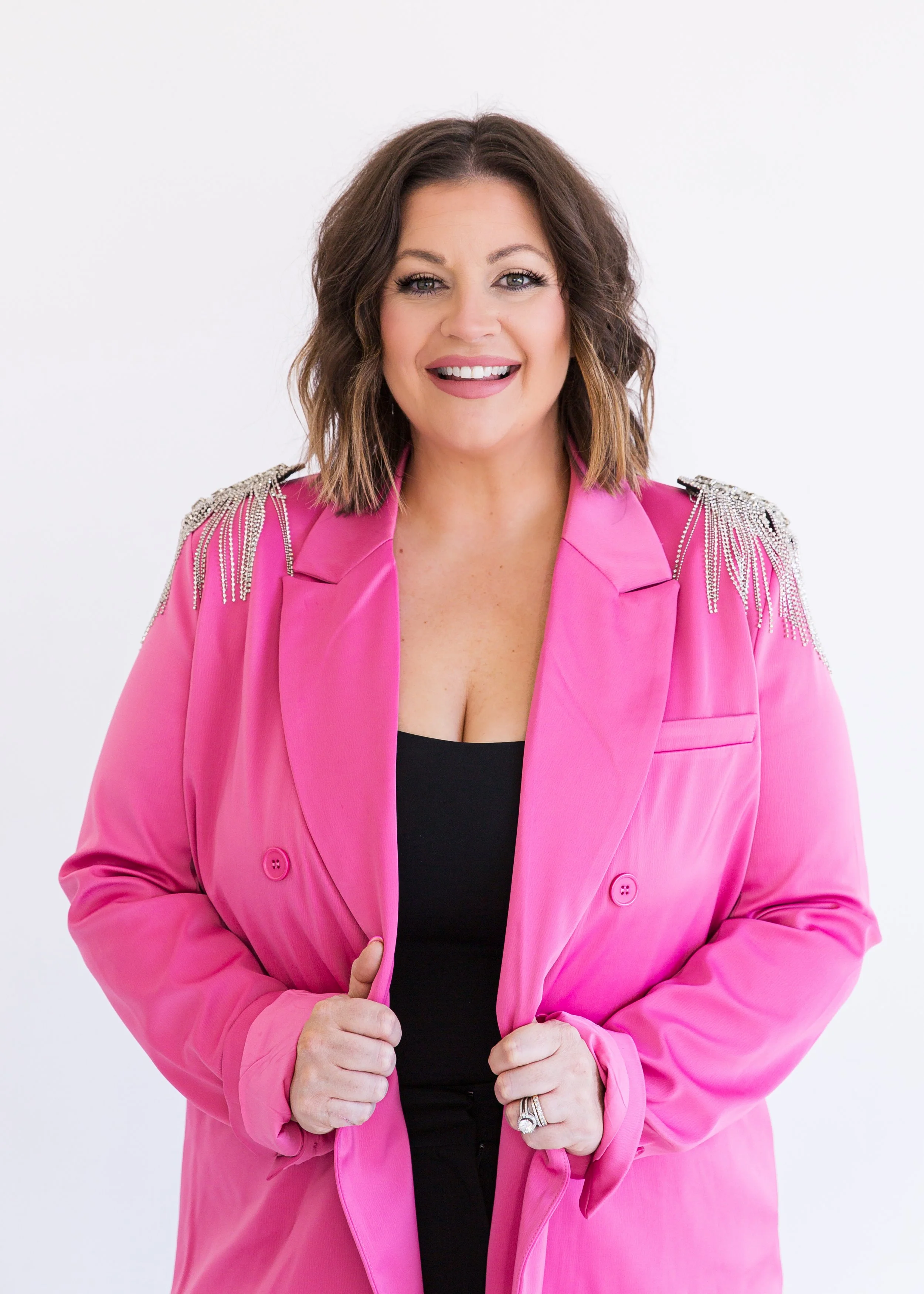 Smiling woman with shoulder-length brown hair wearing a bright pink blazer with rhinestone shoulder embellishments over a black top, holding the lapels of her blazer, against a plain white background.